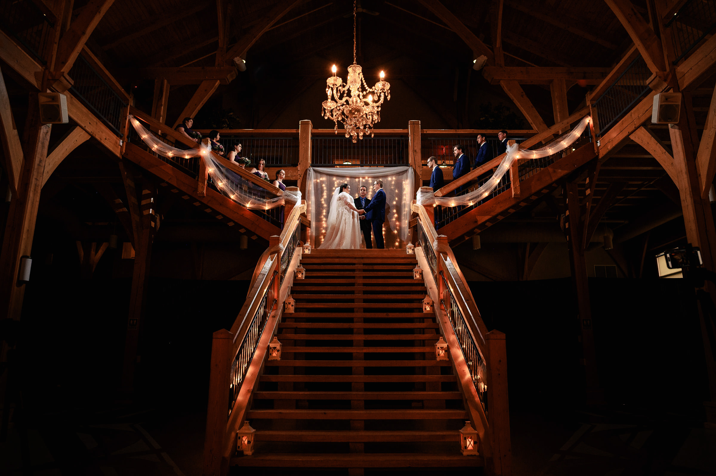 Bride and groom stand atop a grand, dimly lit wooden staircase under a chandelier.