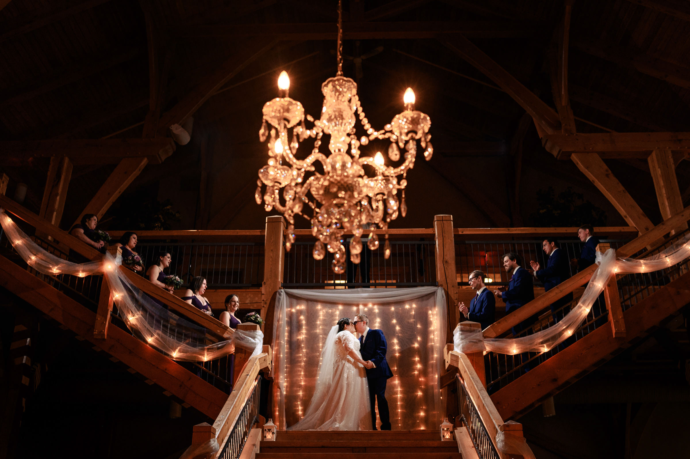 Bride and groom kiss under a chandelier, surrounded by guests on a decorated staircase.