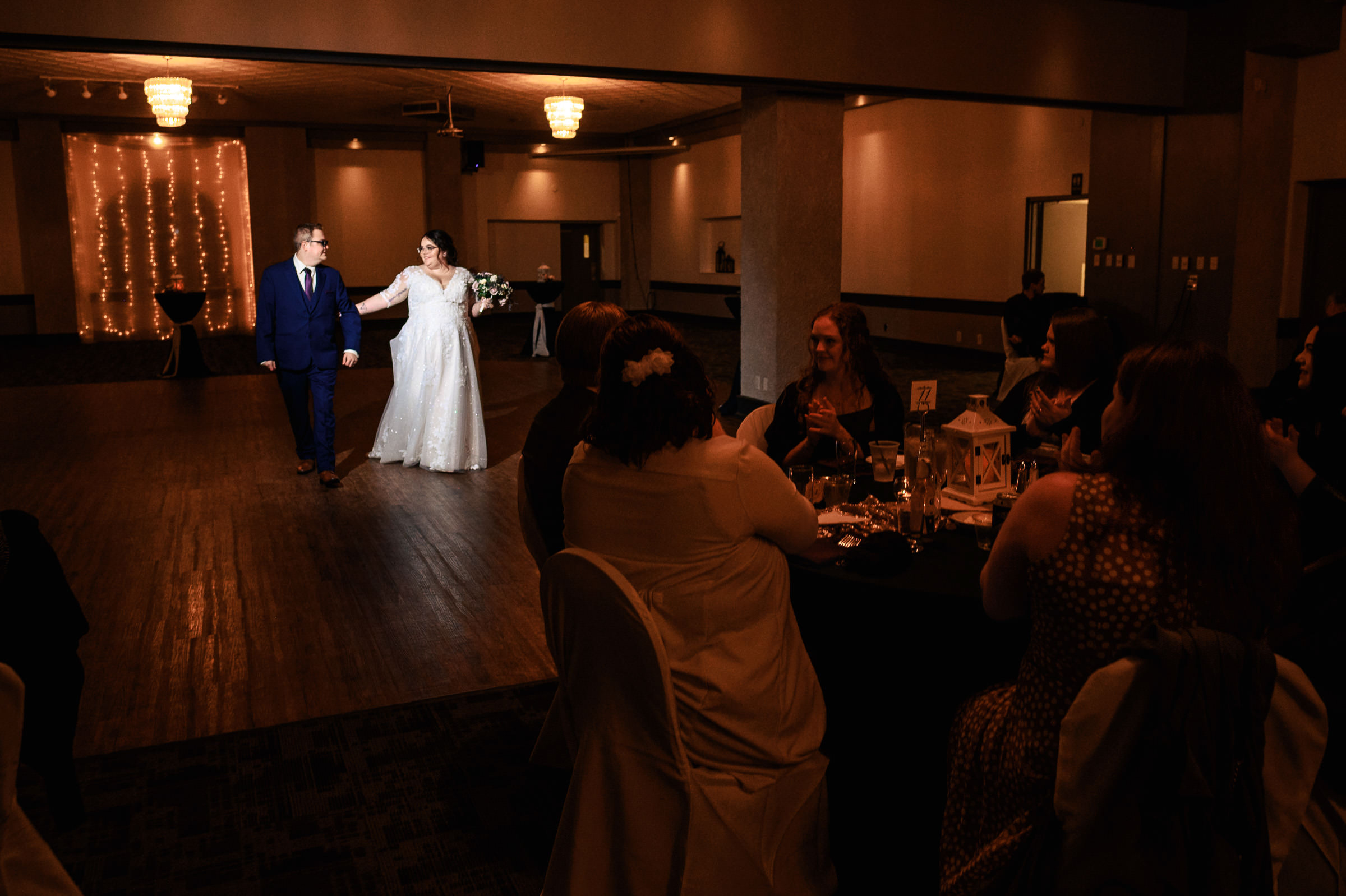 Bride and groom enter a dimly lit venue while guests watch from seated tables.