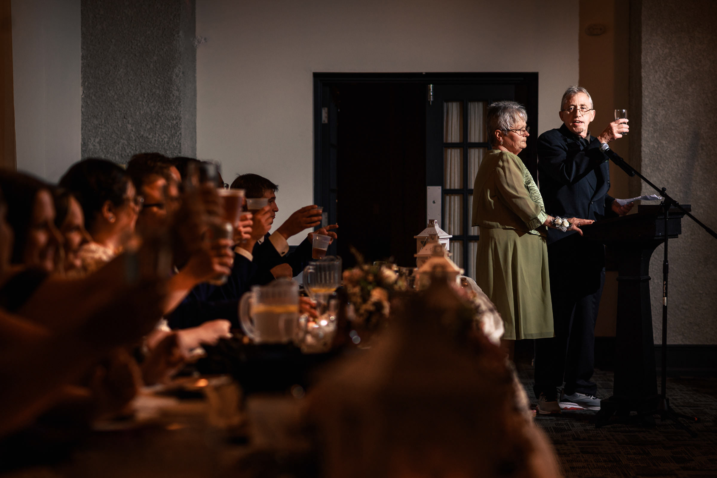 An elderly couple gives a speech at a wedding reception; guests raise glasses in a toast.