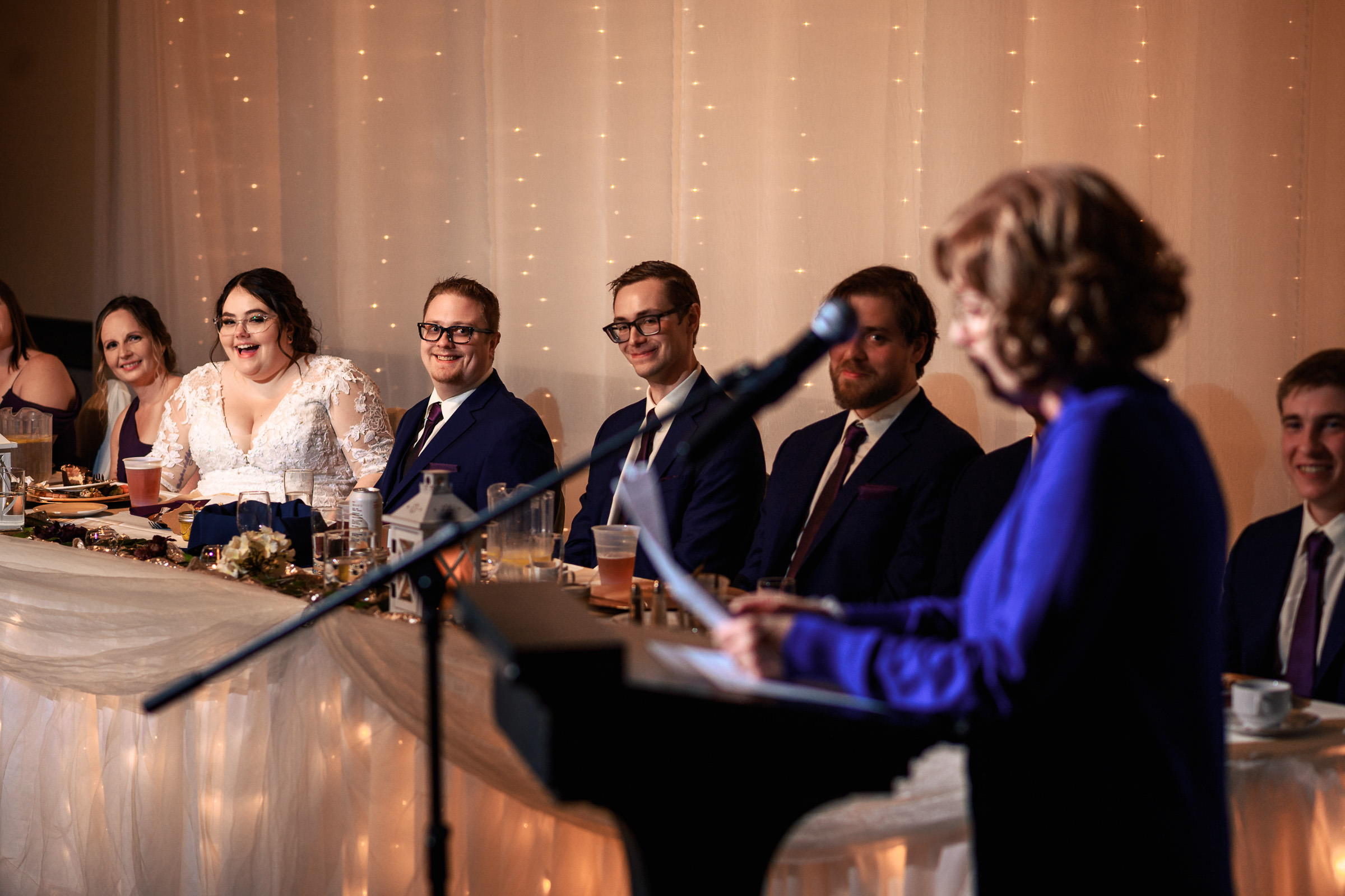 Wedding reception with a speech underway; guests seated at the head table, smiling and attentive.