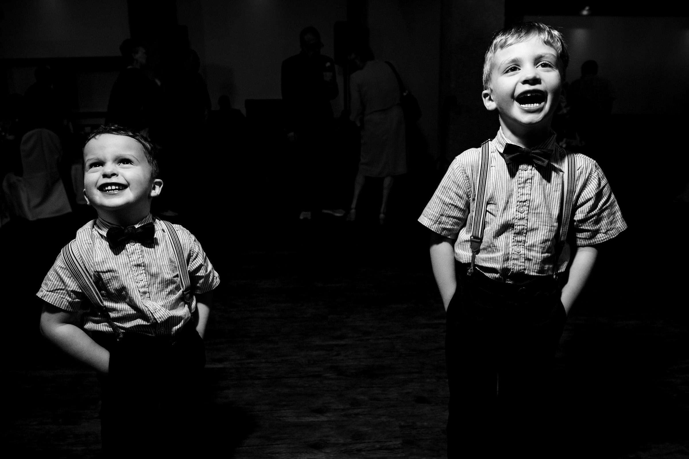 Two young boys in bow ties and suspenders smile widely, standing in a dimly lit room.