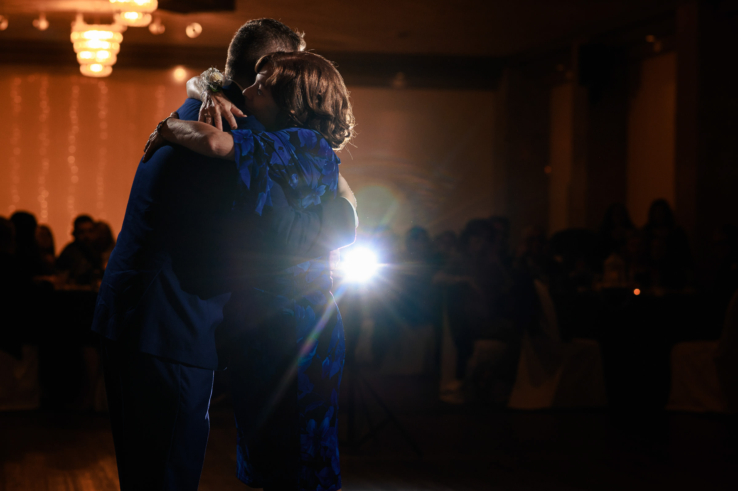 A couple embraces on a dimly lit dance floor with a bright spotlight behind them.