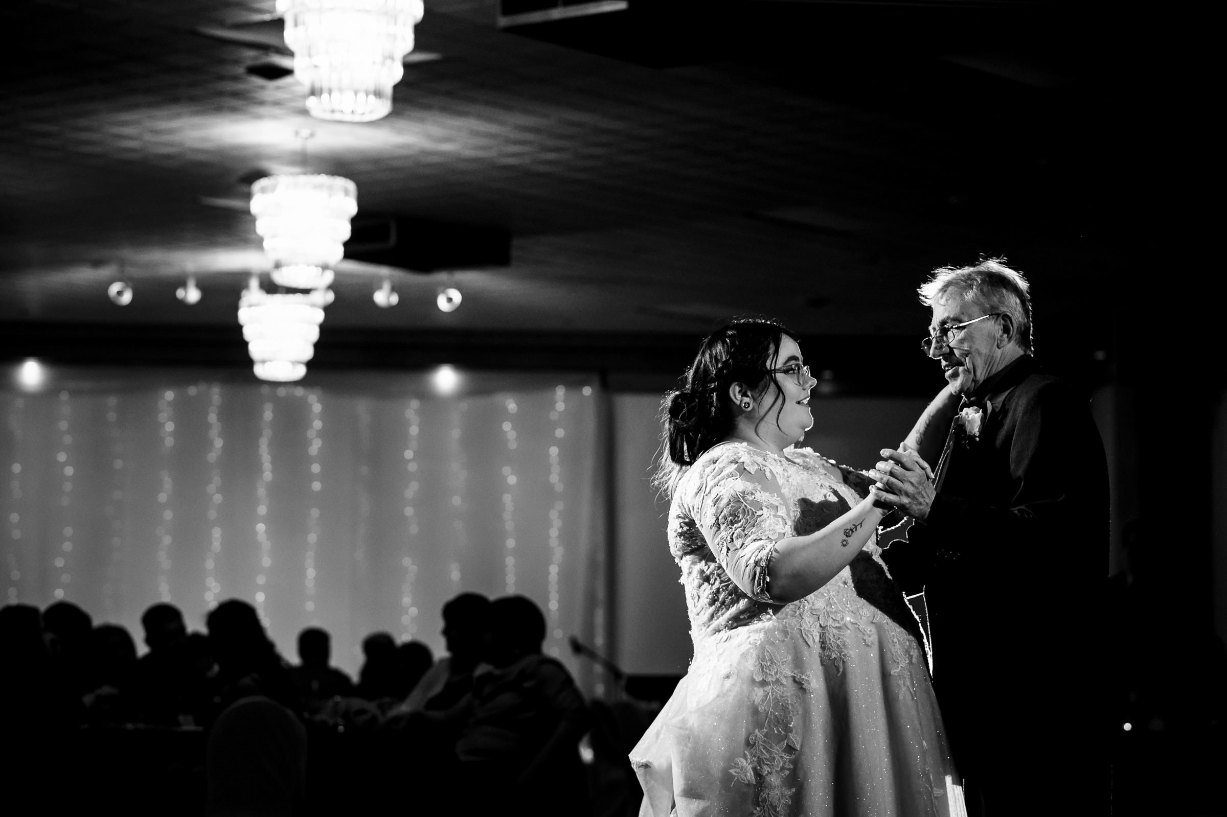 Bride and older man dancing in a dimly lit room with chandeliers.