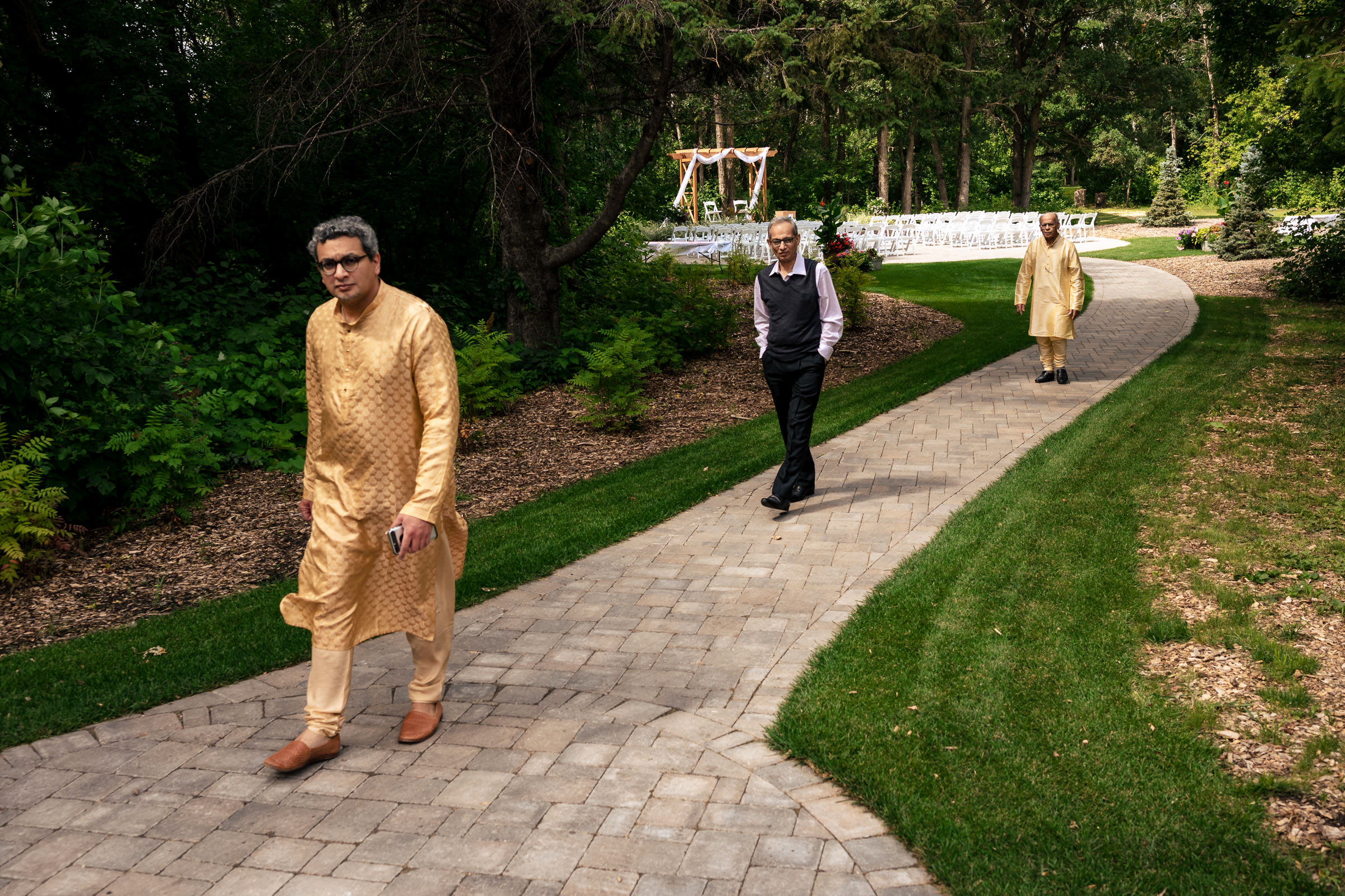 Three people stroll a garden path, with wedding seating and canopy in the background.