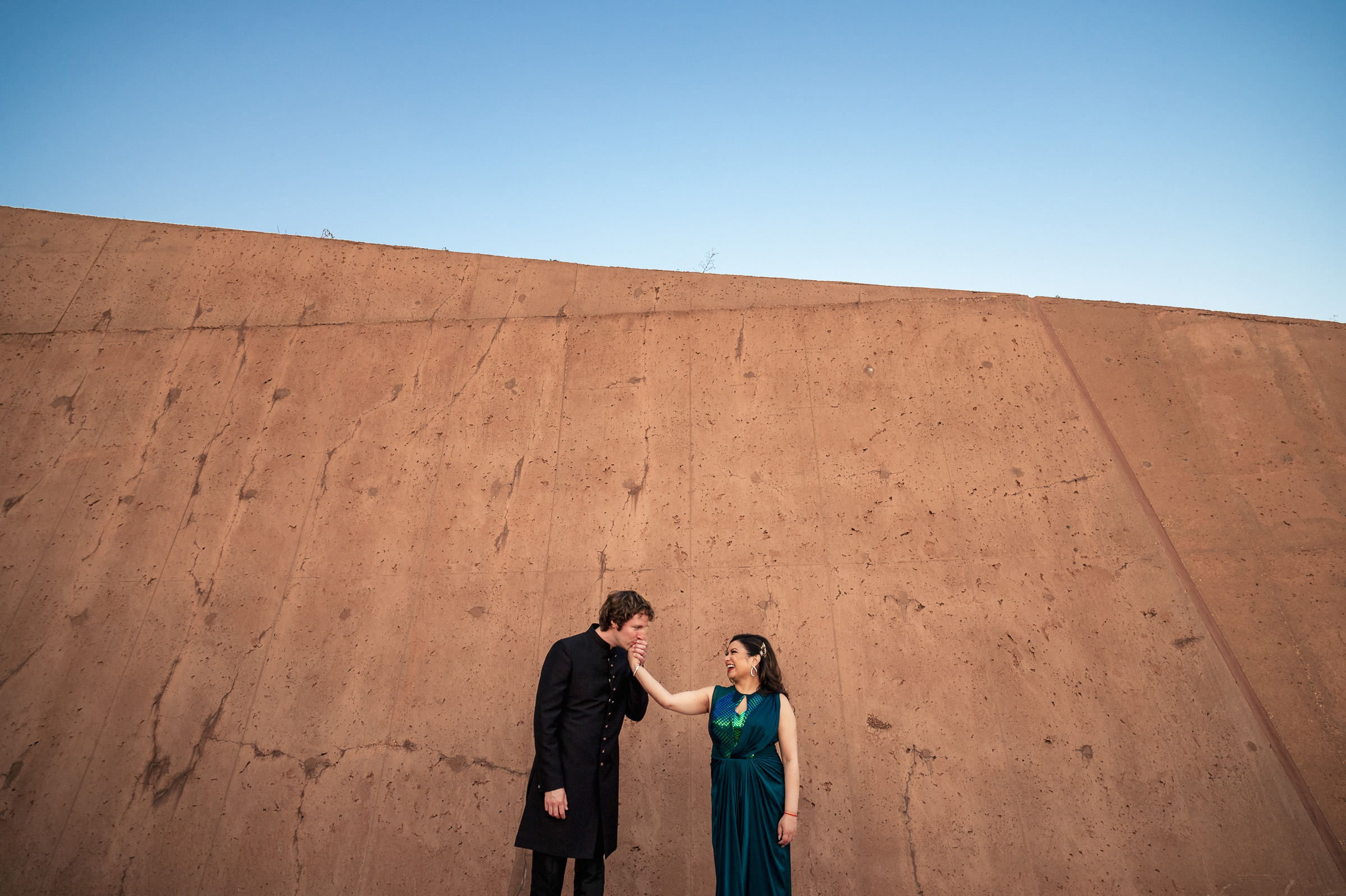 Two people stand against a large, textured concrete wall under Winnipeg's clear blue sky.