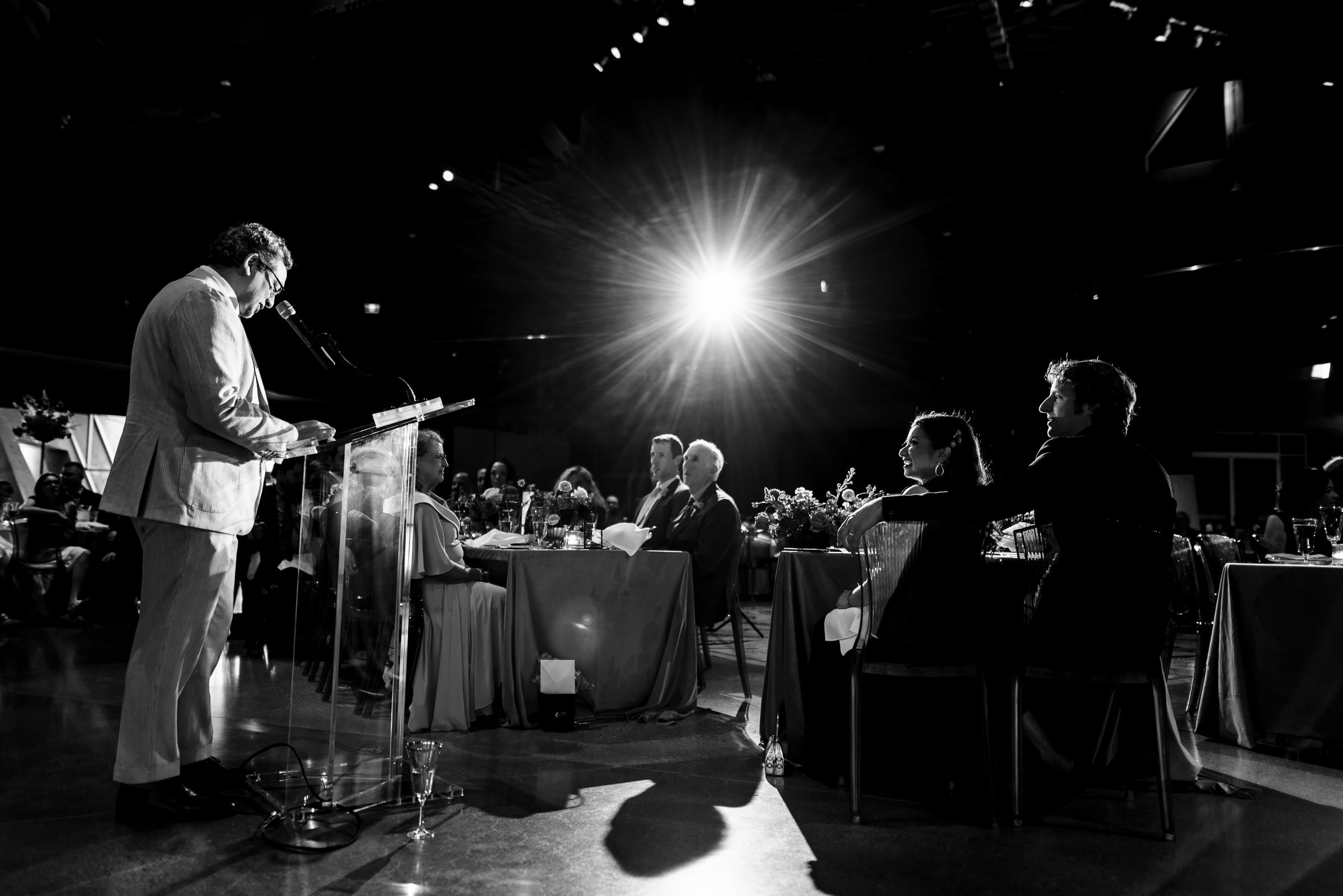 Man in suit speaks at podium during a Winnipeg wedding, spotlight overhead in dimly lit room.