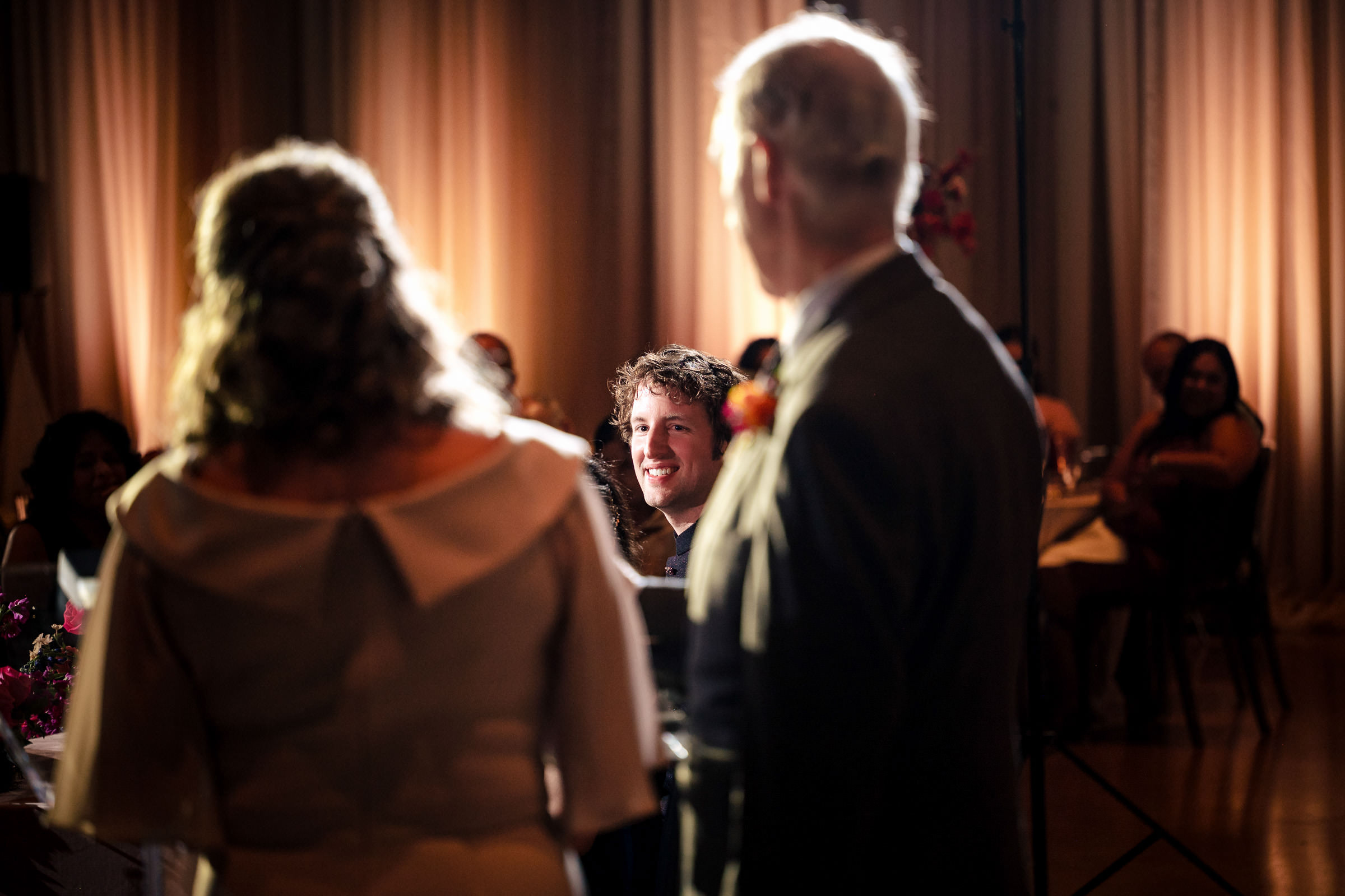 A smiling person sits at a table, watching two people in focus at a dimly lit Winnipeg wedding.