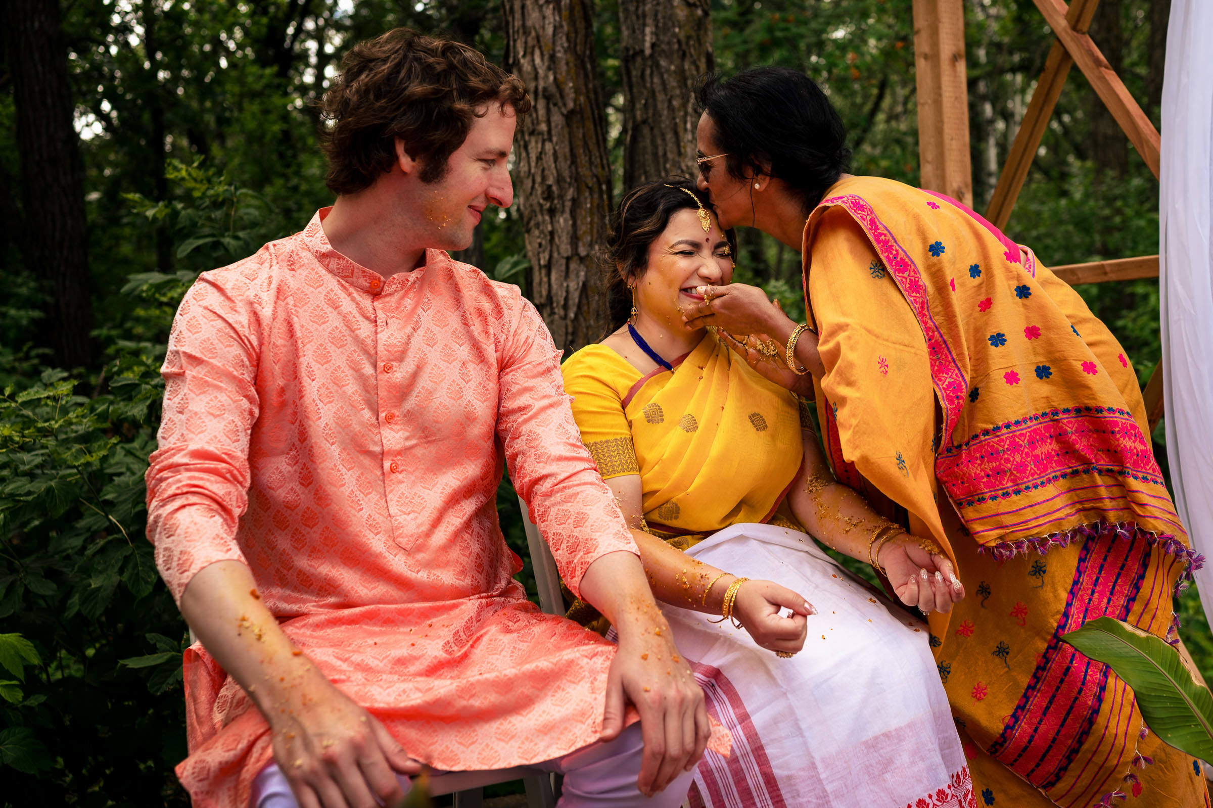 Three people smiling at a Winnipeg wedding, in colorful traditional attire, one feeding another.