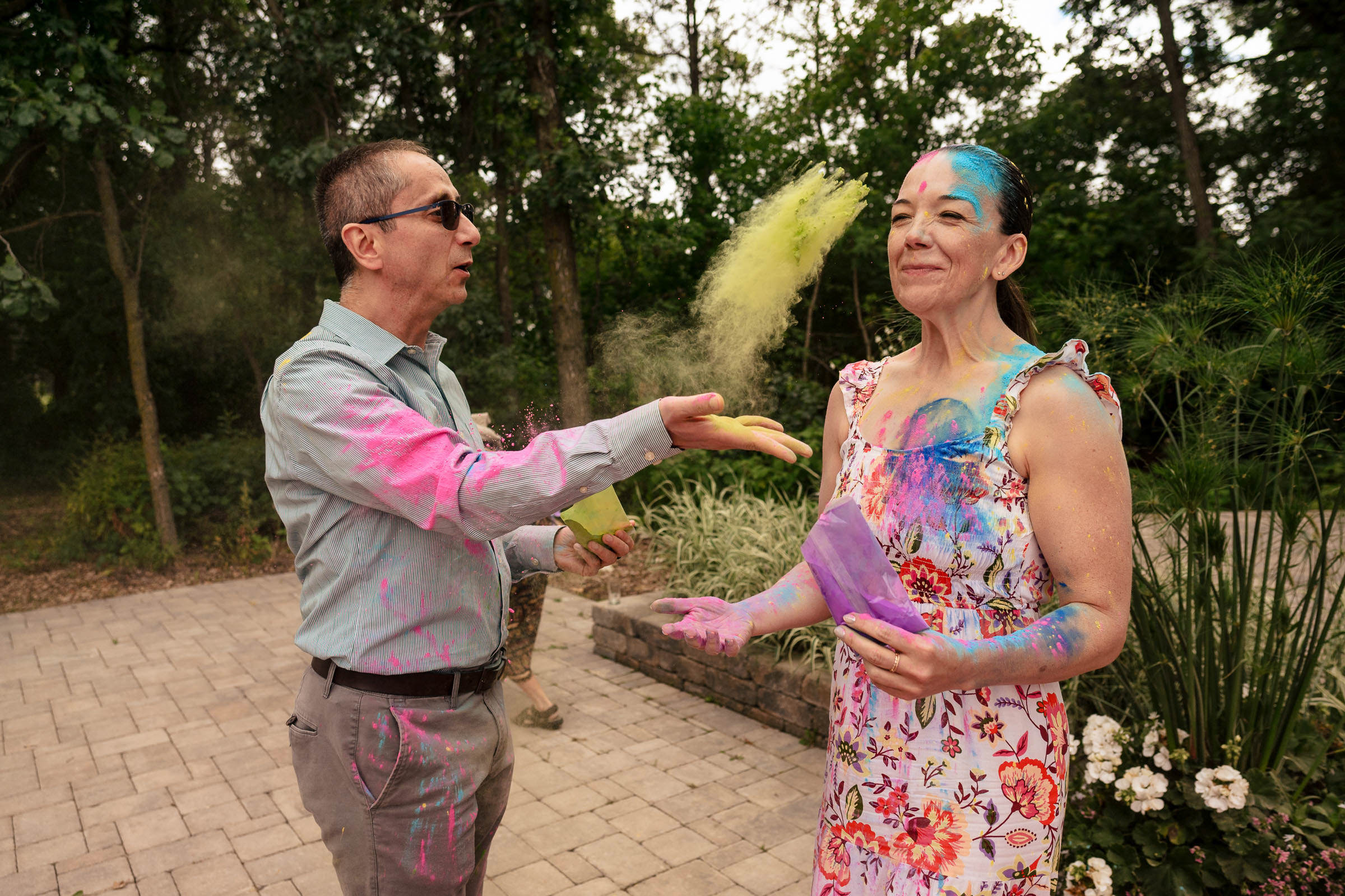 Two people in a garden, like a Winnipeg wedding, throw colored powder and vibrant hues onto each other.