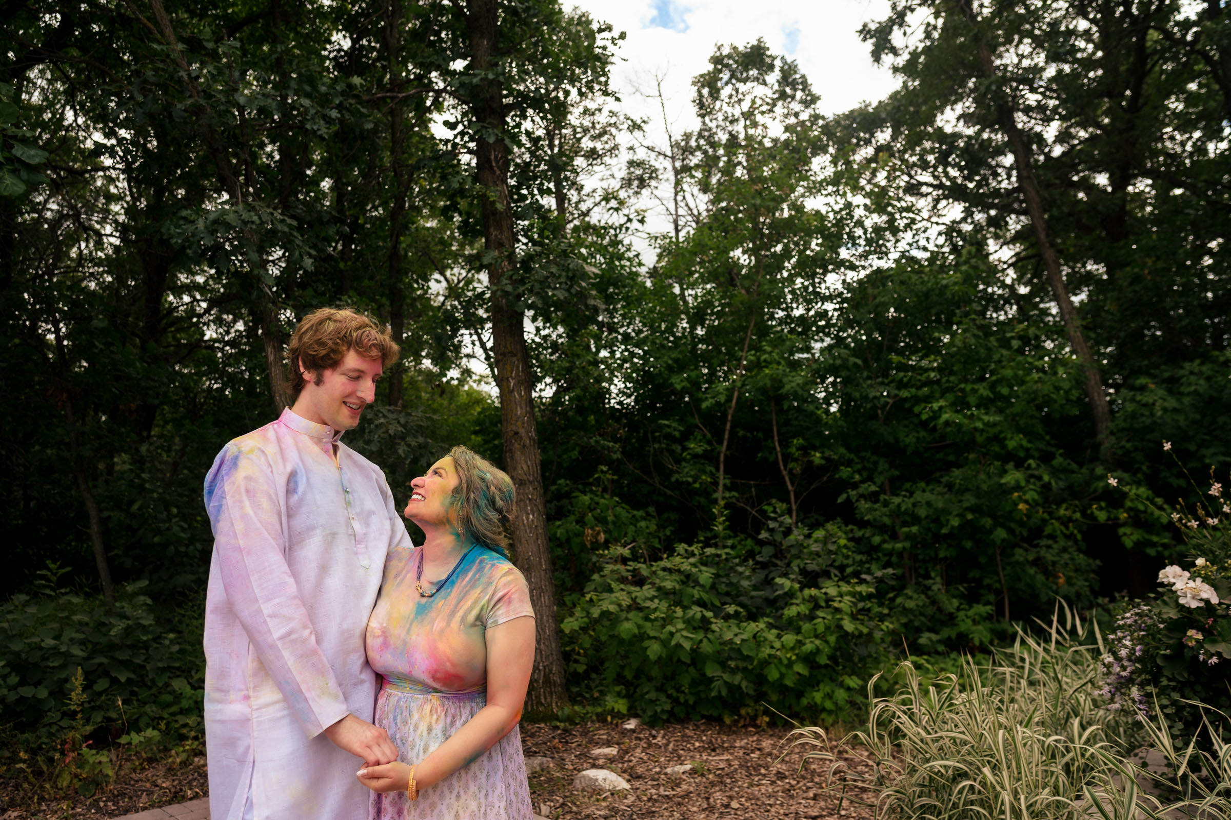 Couple in colorful attire stands in a wooded area, smiling at each other during their Winnipeg wedding.
