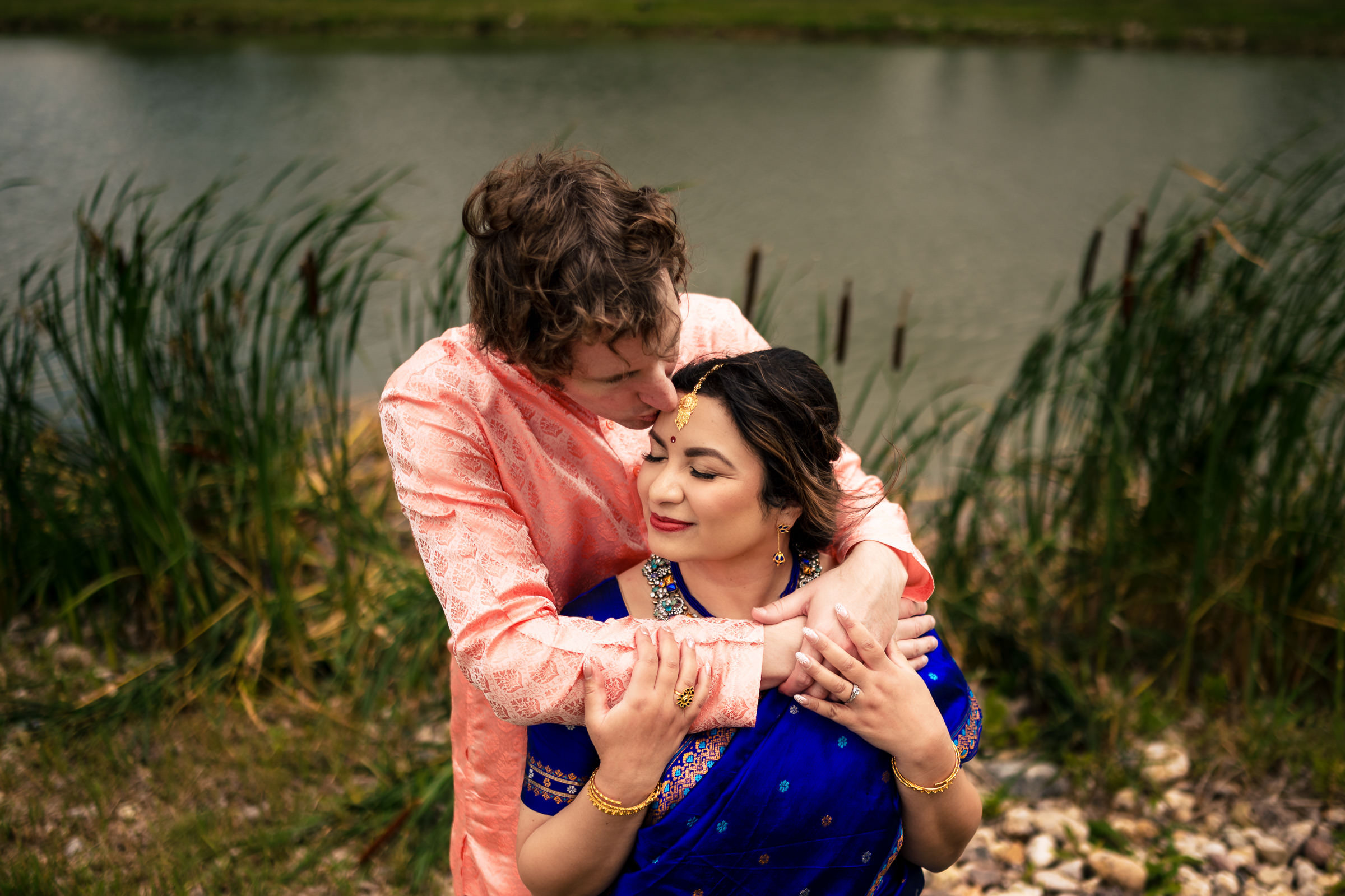 Couple embracing by a pond; she in blue sari, he in peach kurta at a Winnipeg wedding.