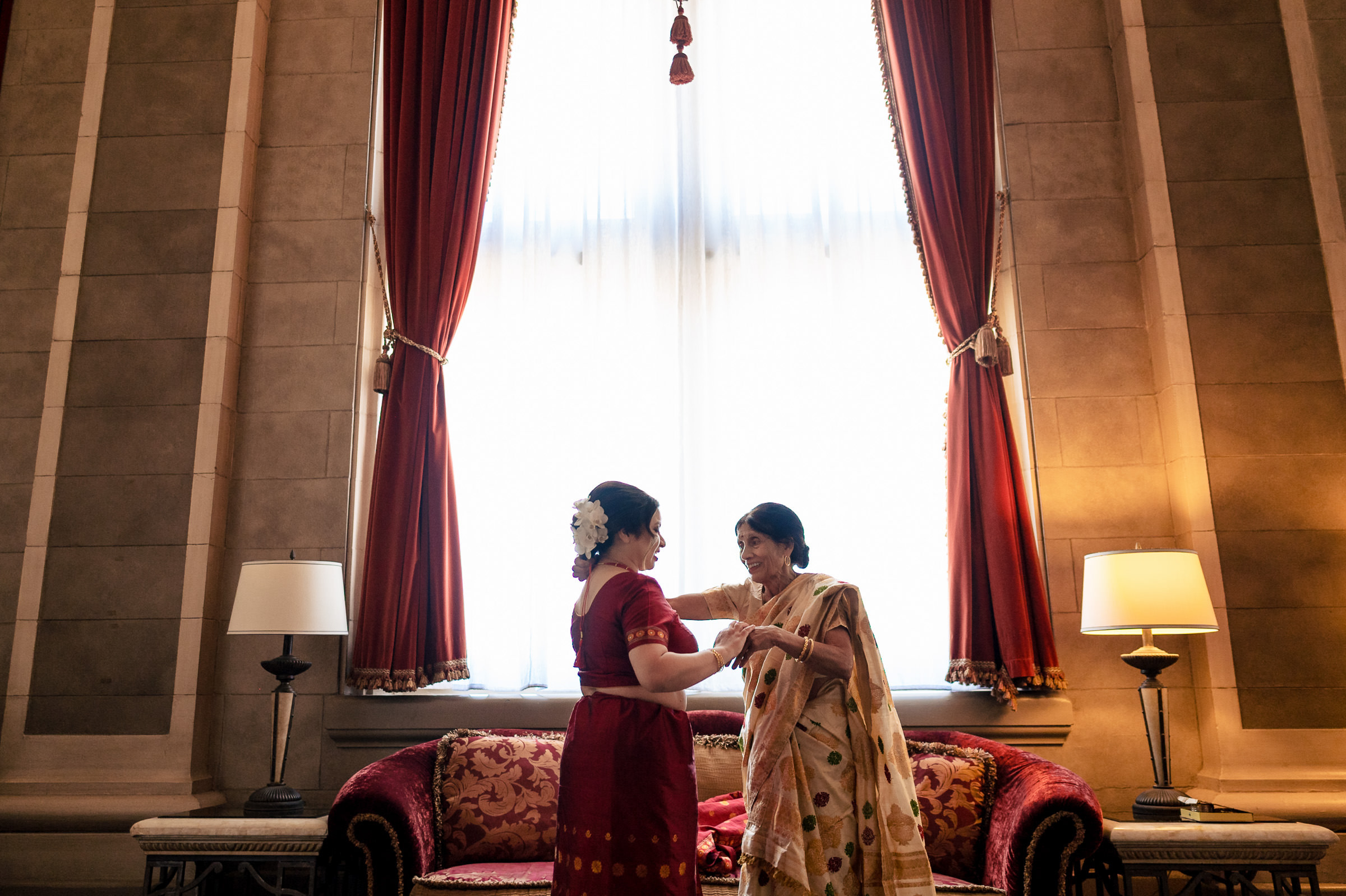 Two women in traditional attire grace a Winnipeg wedding near red-curtained sofas.