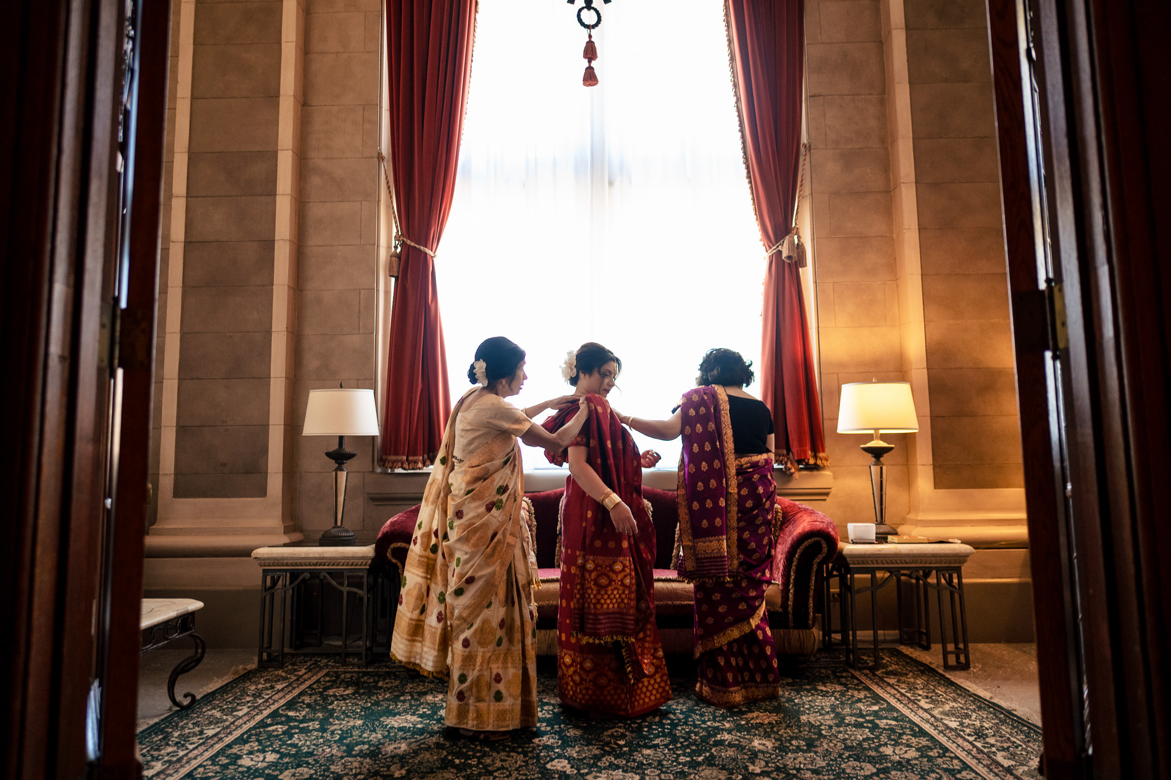 Three women in sarees gather near a sofa at a Winnipeg wedding, framed by tall red curtains.
