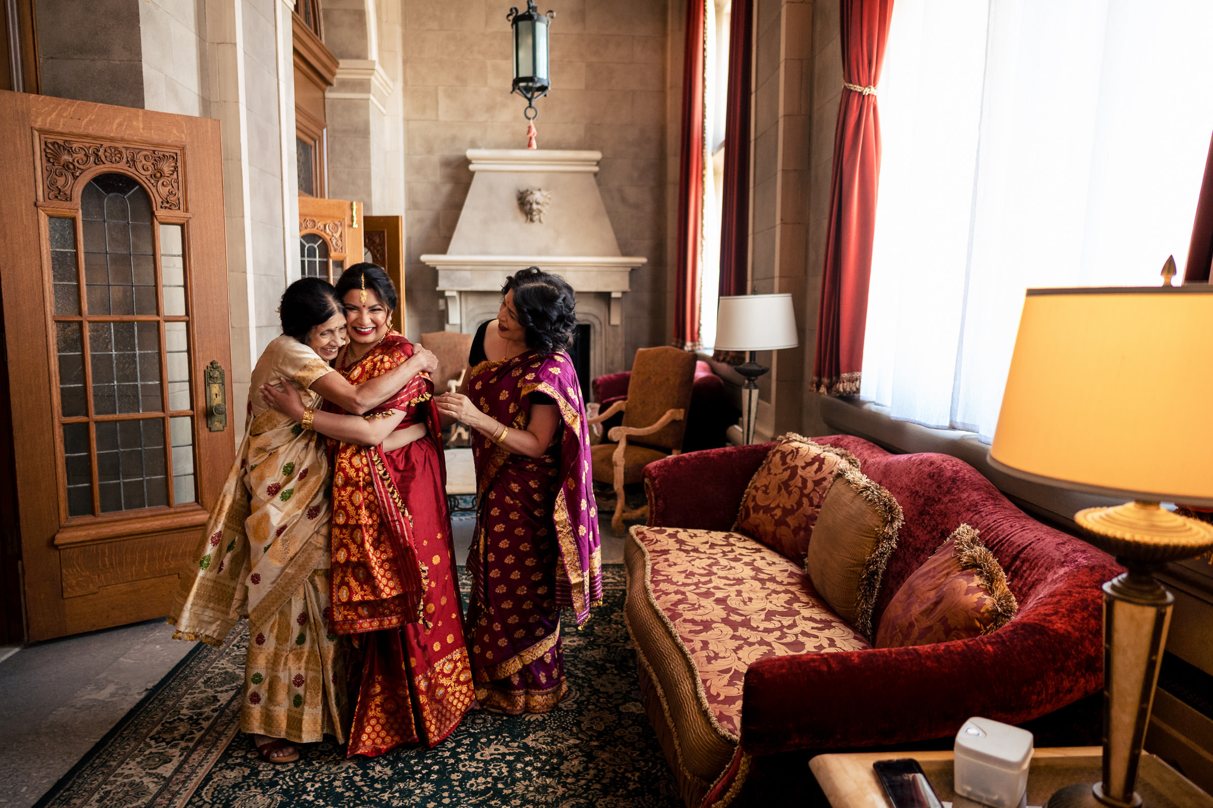 Three women in traditional attire hug joyfully at a Winnipeg wedding by a cozy fireplace.