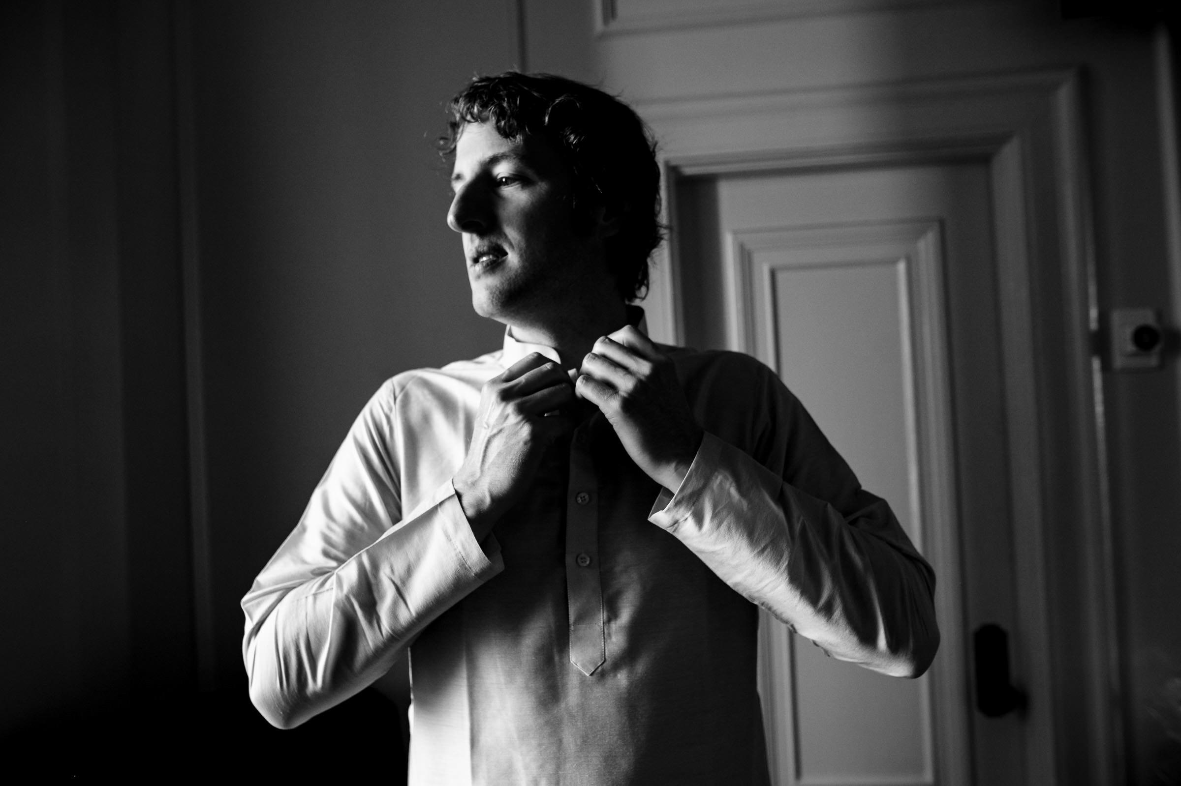 Man in a white shirt adjusts his collar before the Winnipeg wedding in a dimly lit room.