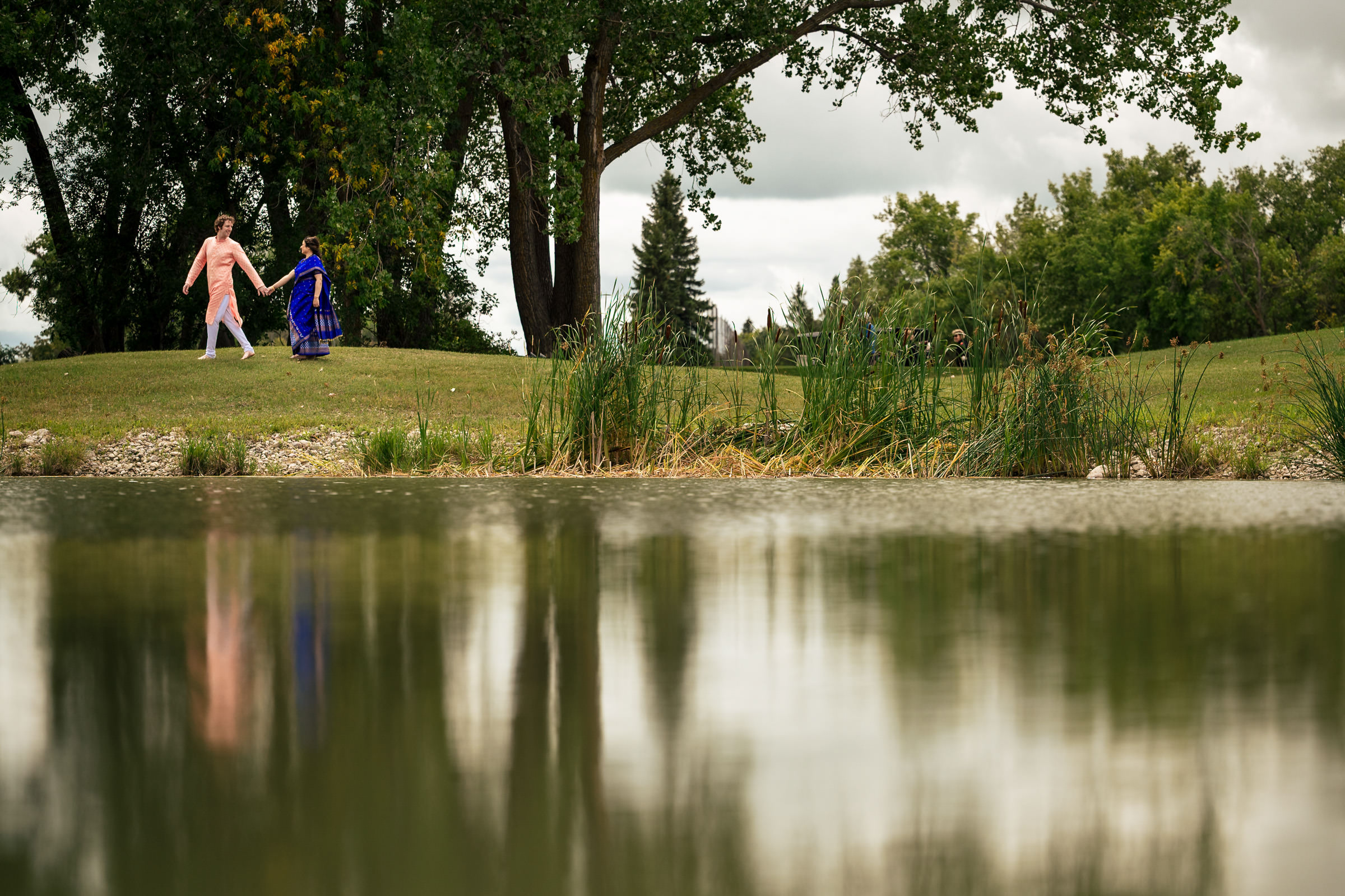 A Winnipeg wedding couple strolls hand in hand by a tranquil pond with trees and grass.