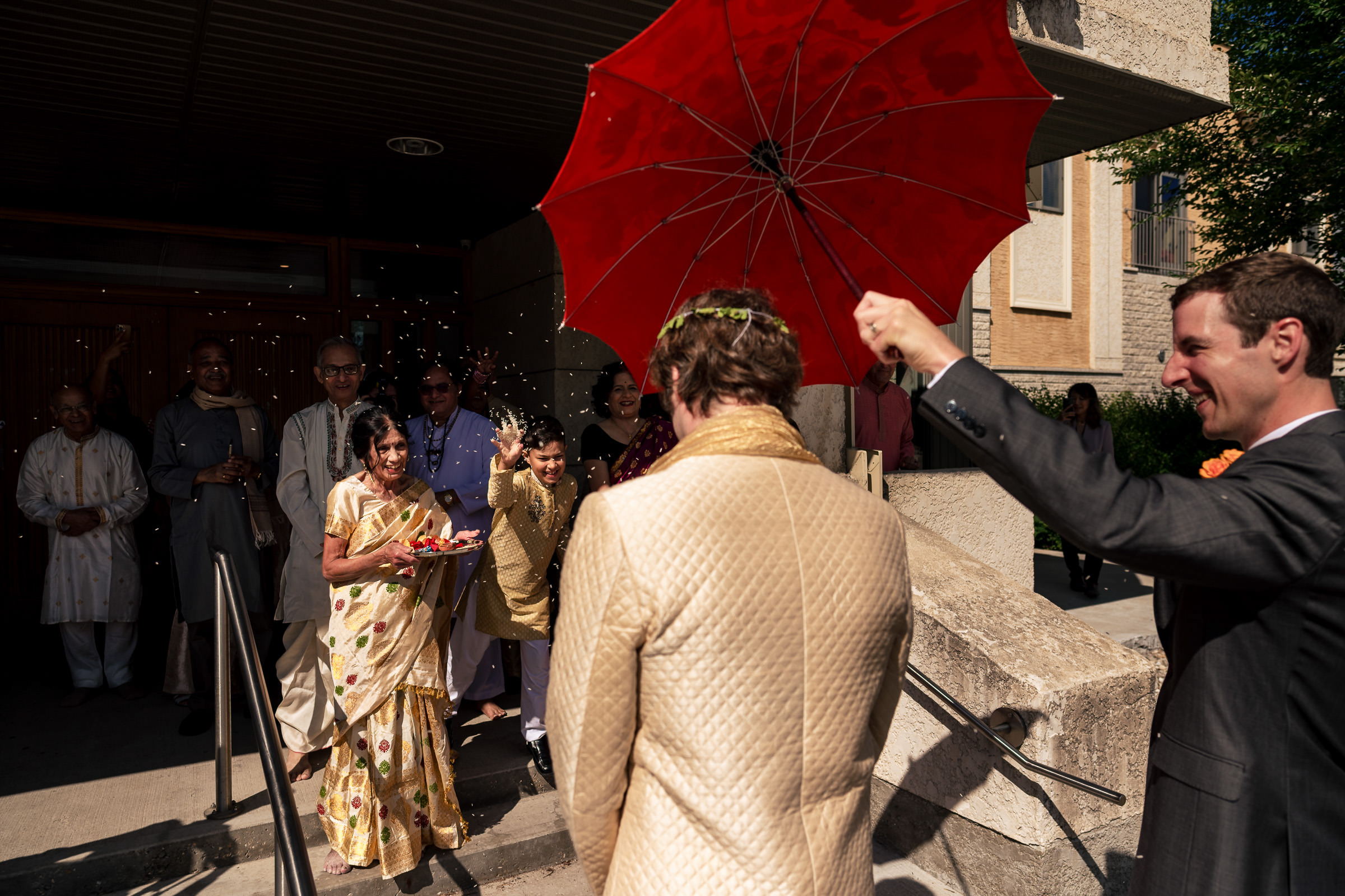 Guests greet a couple outside a building during a Winnipeg wedding, with a red umbrella overhead.