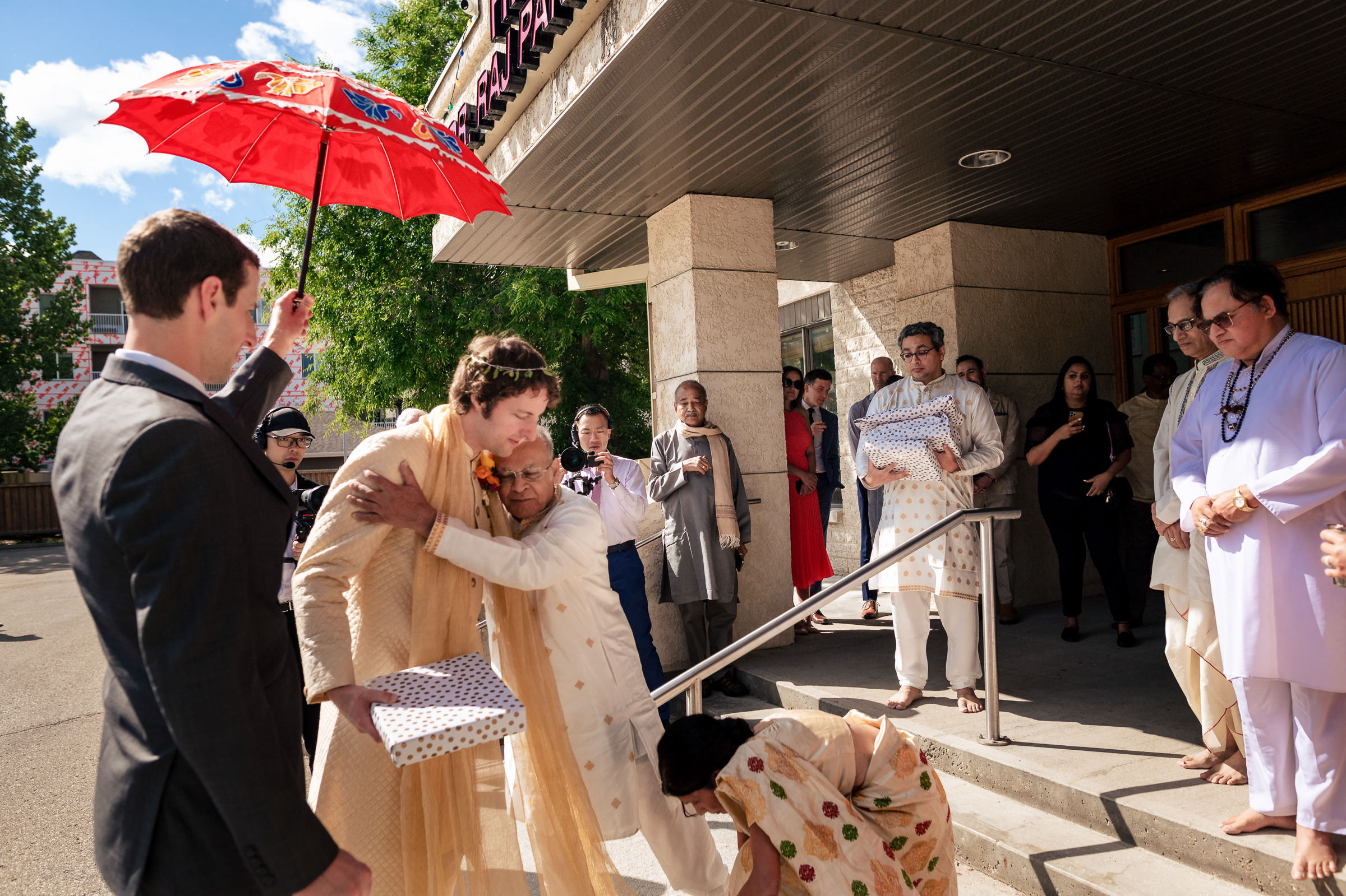 Person in traditional attire hugs another at a Winnipeg wedding; guests stand holding gifts.