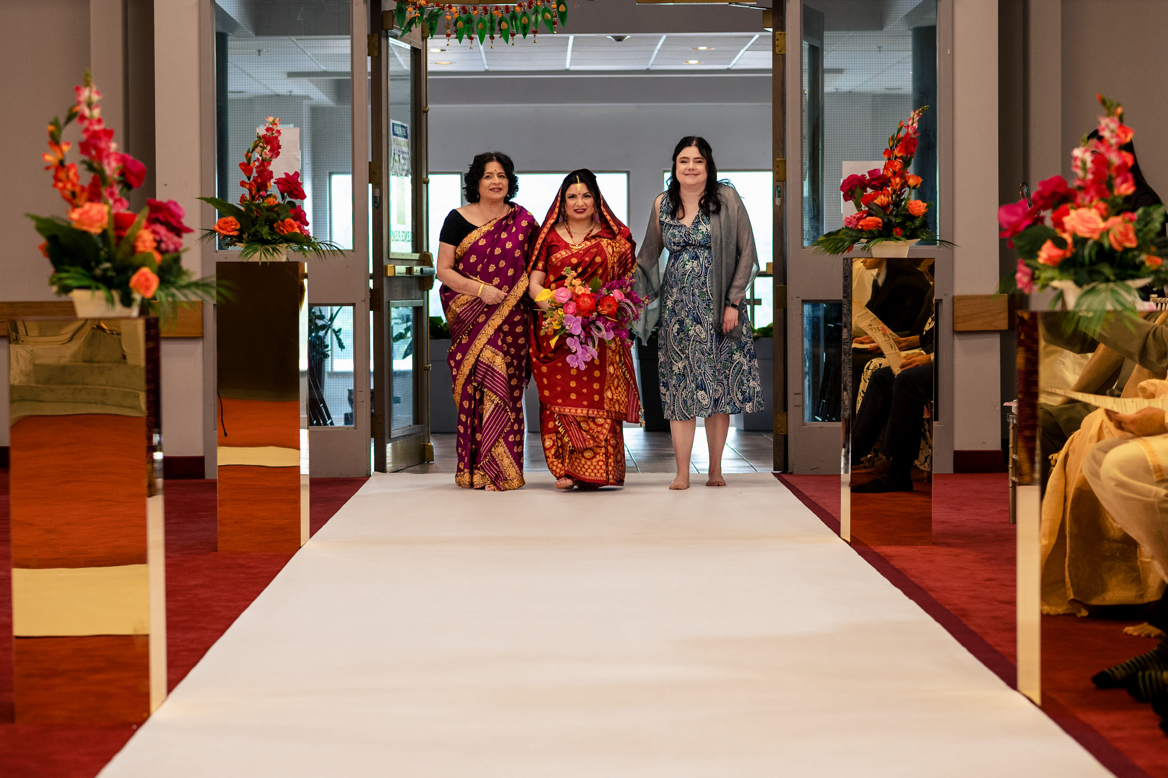 Three women walking down an aisle at a Winnipeg wedding; one in a red sari holds flowers.