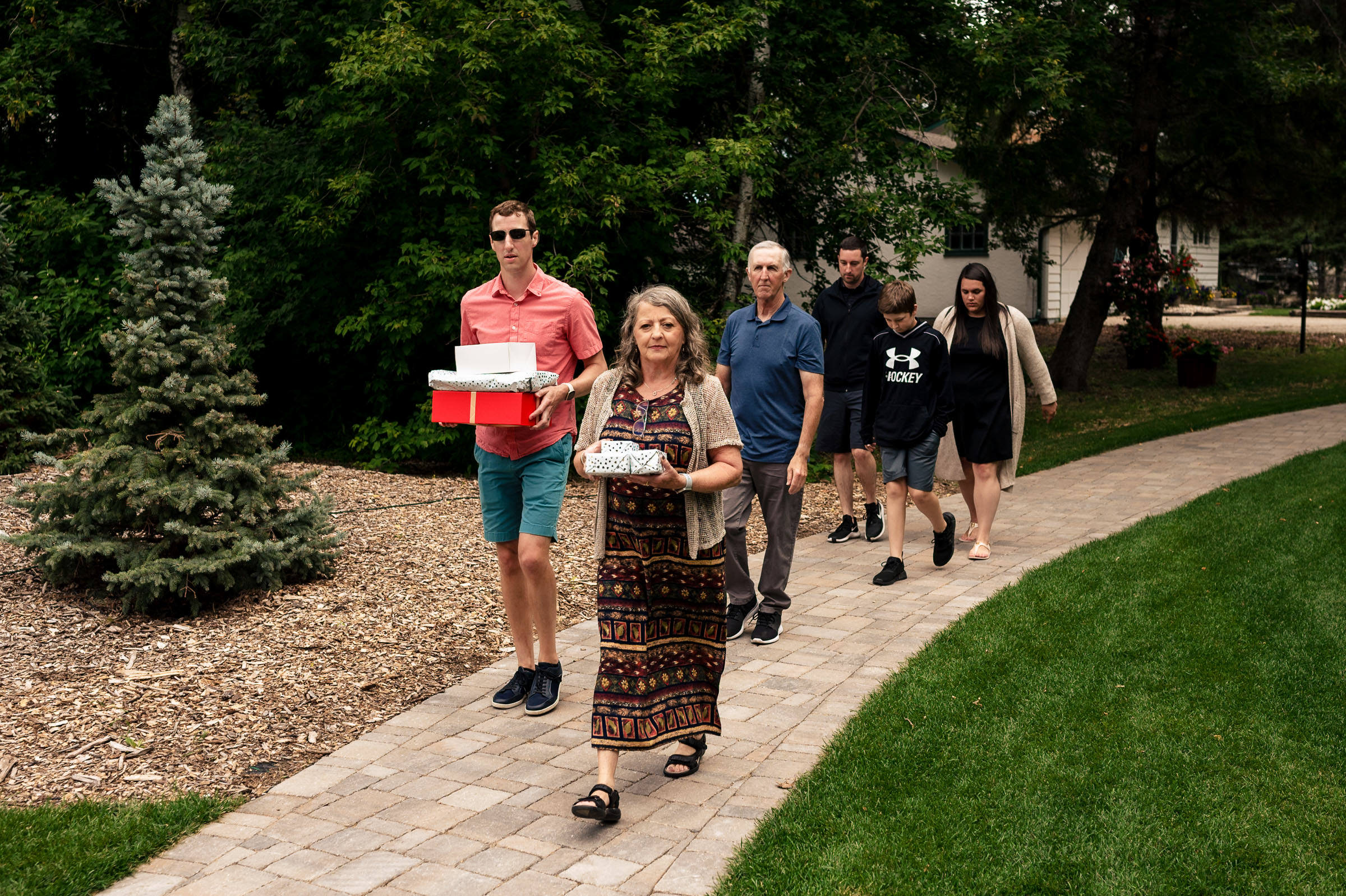 A group strolls down a garden path, carrying boxed items at a Winnipeg wedding.