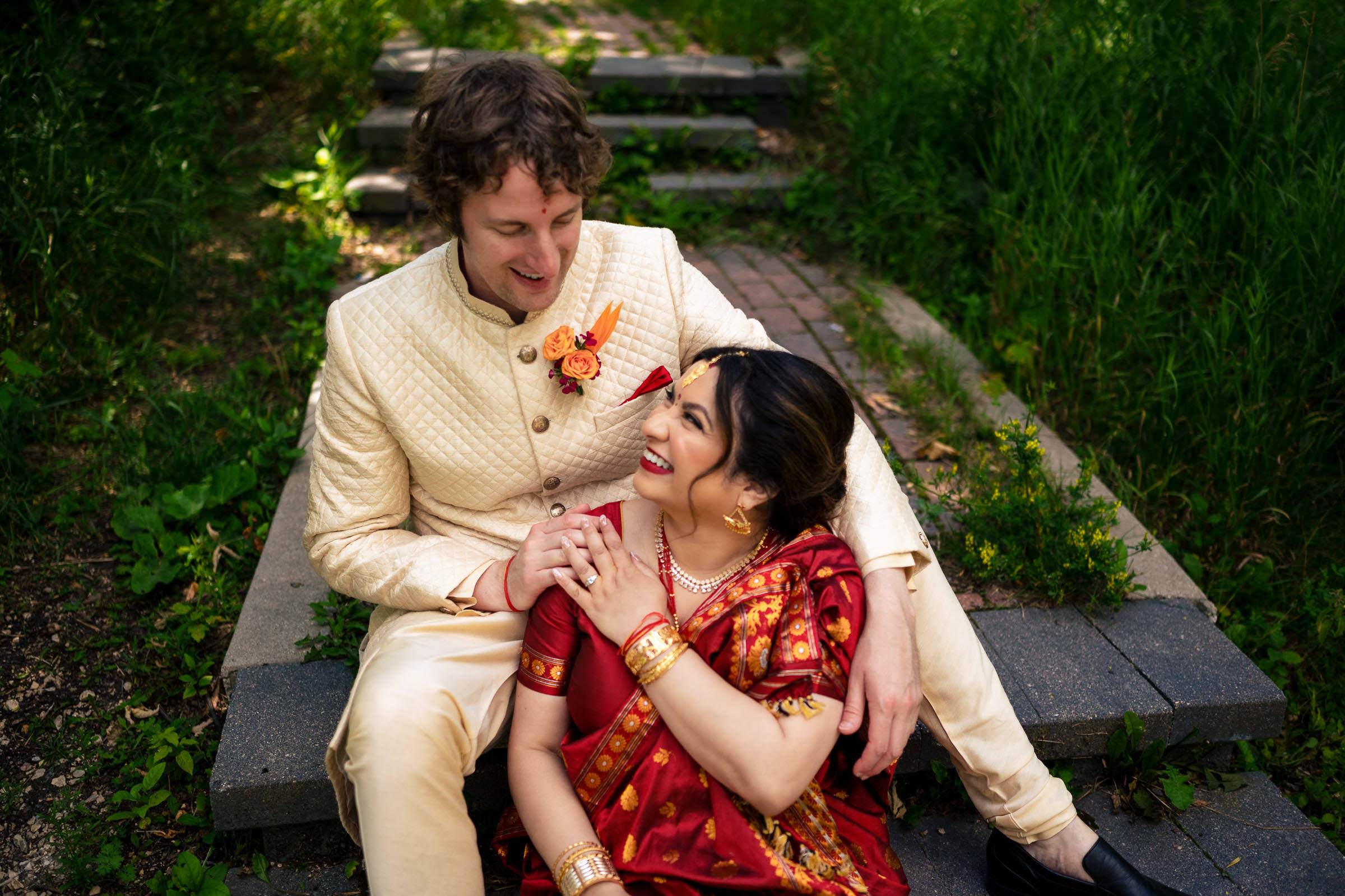 A couple smiles on the steps in traditional attire, surrounded by Winnipeg wedding greenery.
