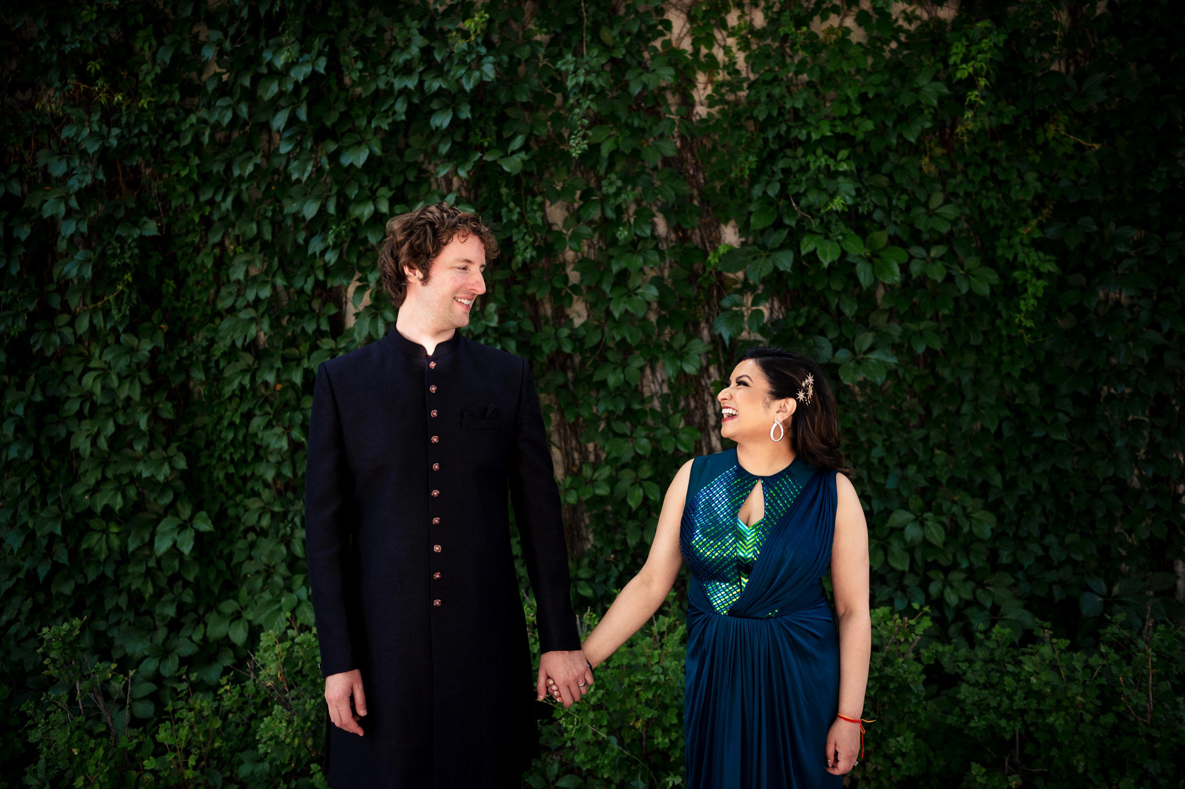 A couple holds hands, smiling in front of a lush green backdrop at their Winnipeg wedding.