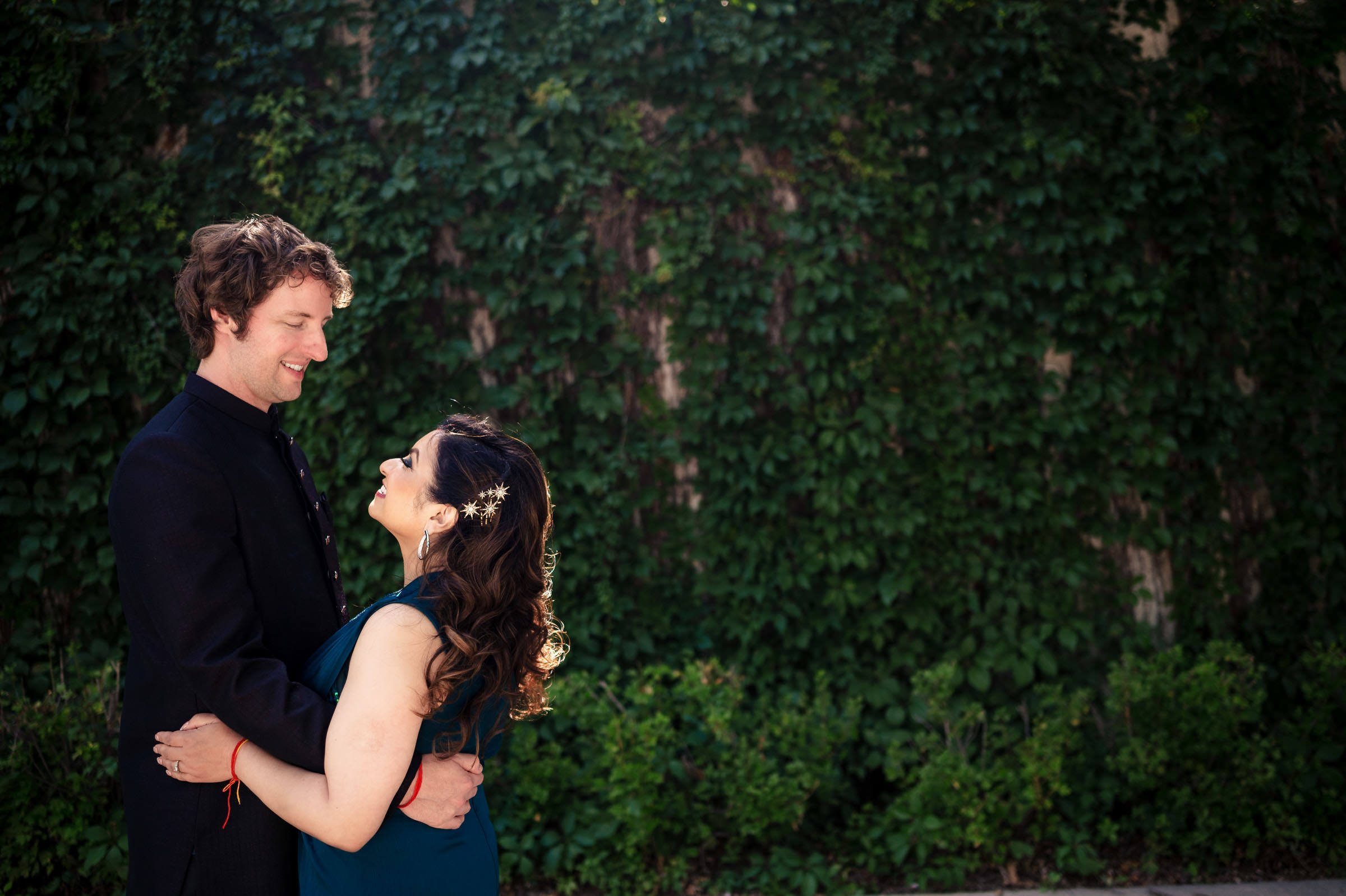 A couple embraces, smiling, in front of a leafy green wall at their Winnipeg wedding.