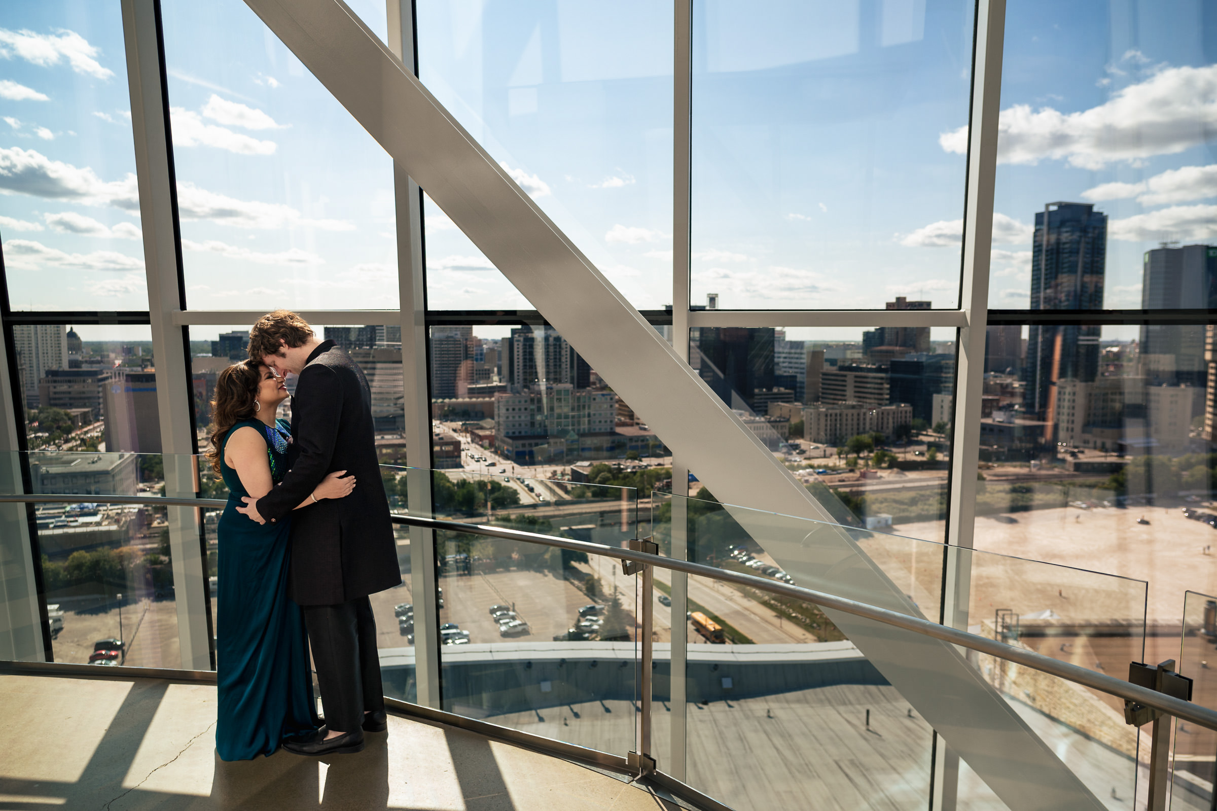 A couple embraces in a glass building with Winnipeg's cityscape in the background.