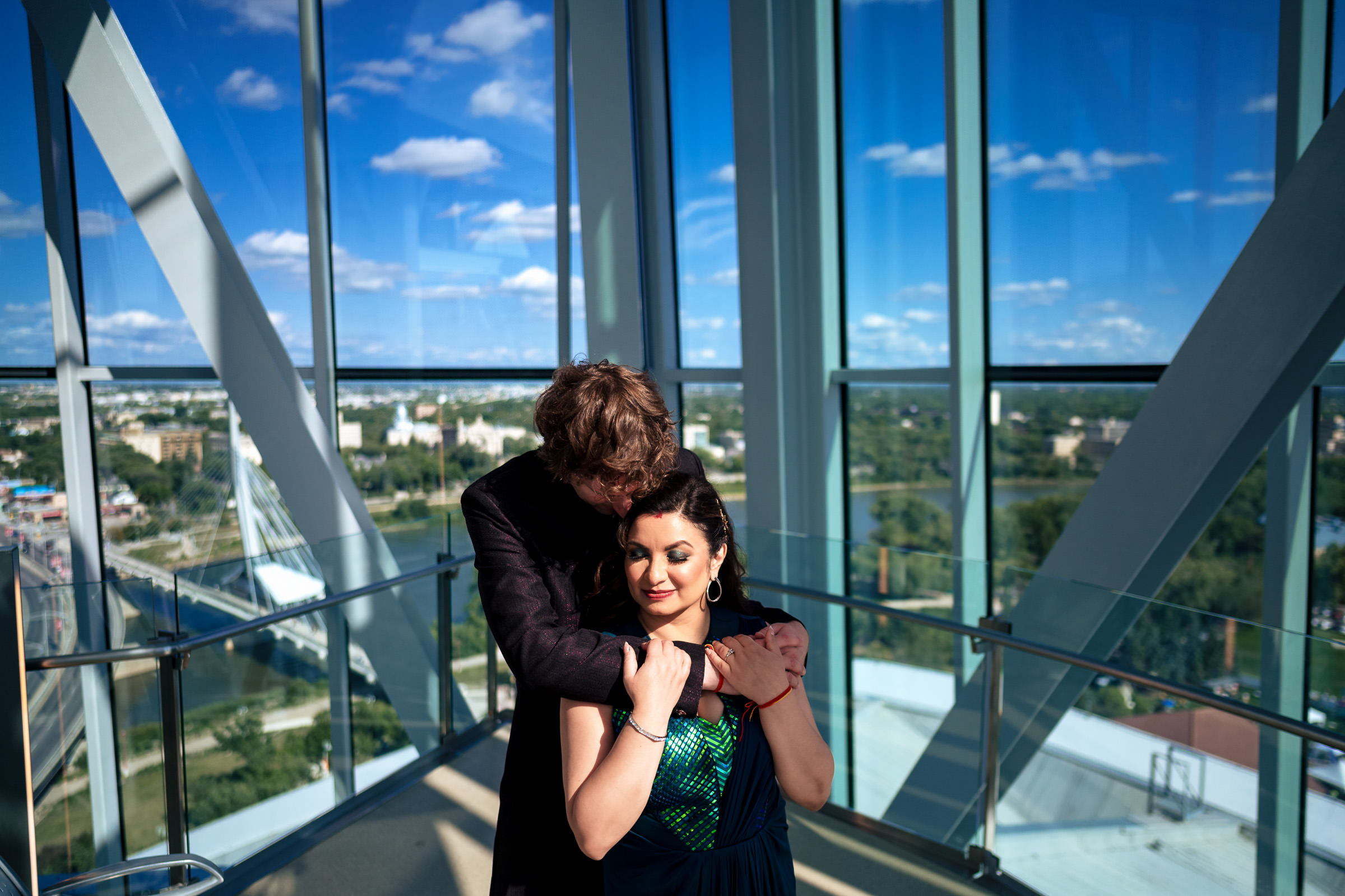Couple on a glass balcony with Winnipeg wedding cityscape and blue sky in the background.