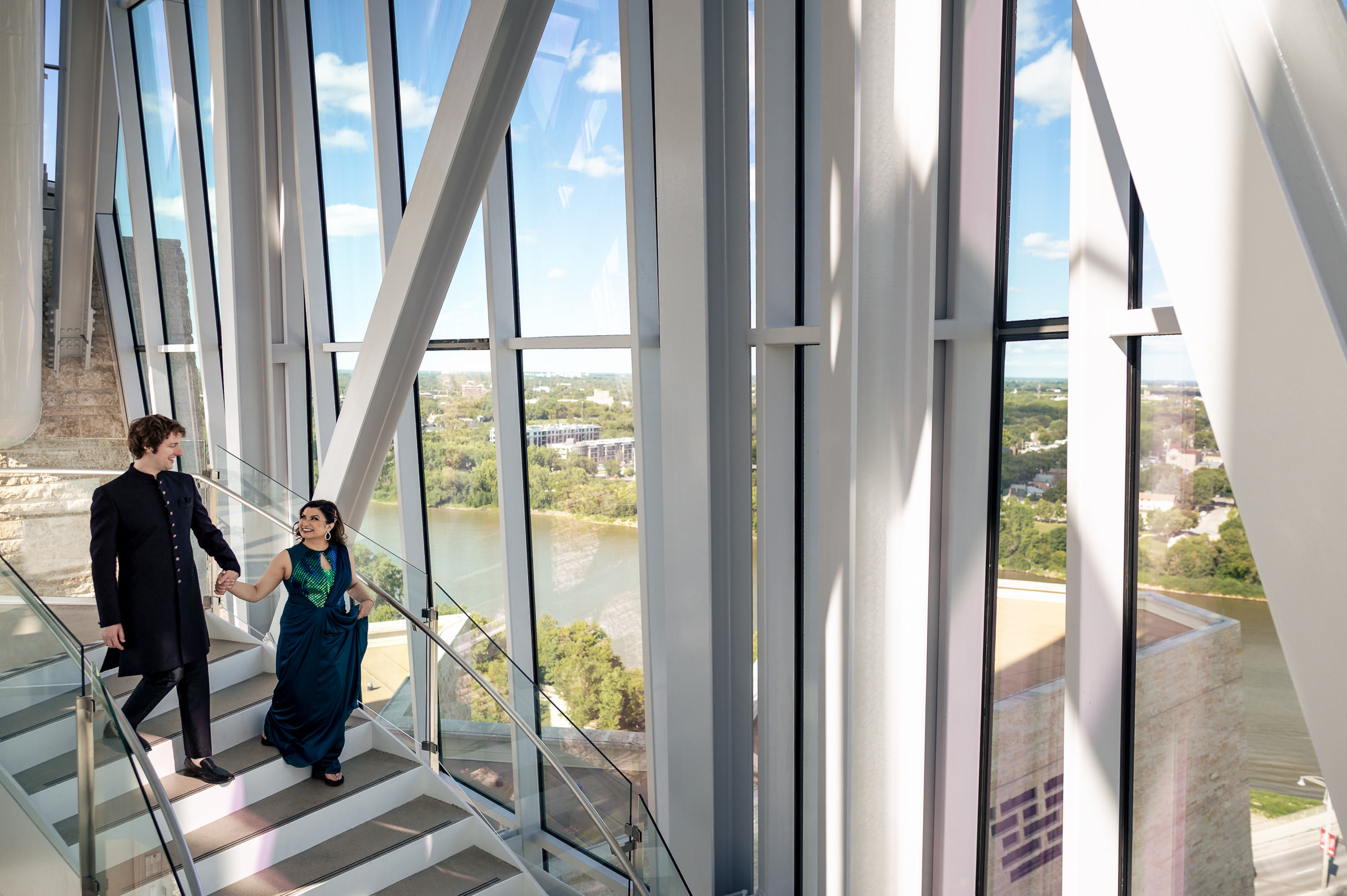 A couple in formal attire ascends a modern glass staircase at their Winnipeg wedding.