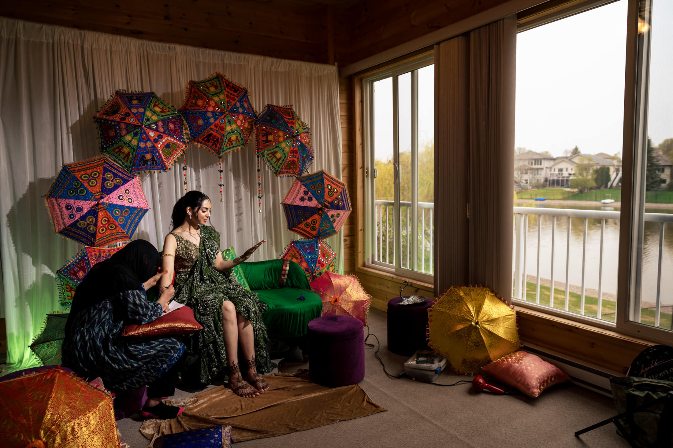 Woman sitting on a sofa with colorful umbrellas, another woman applying henna on her hand.