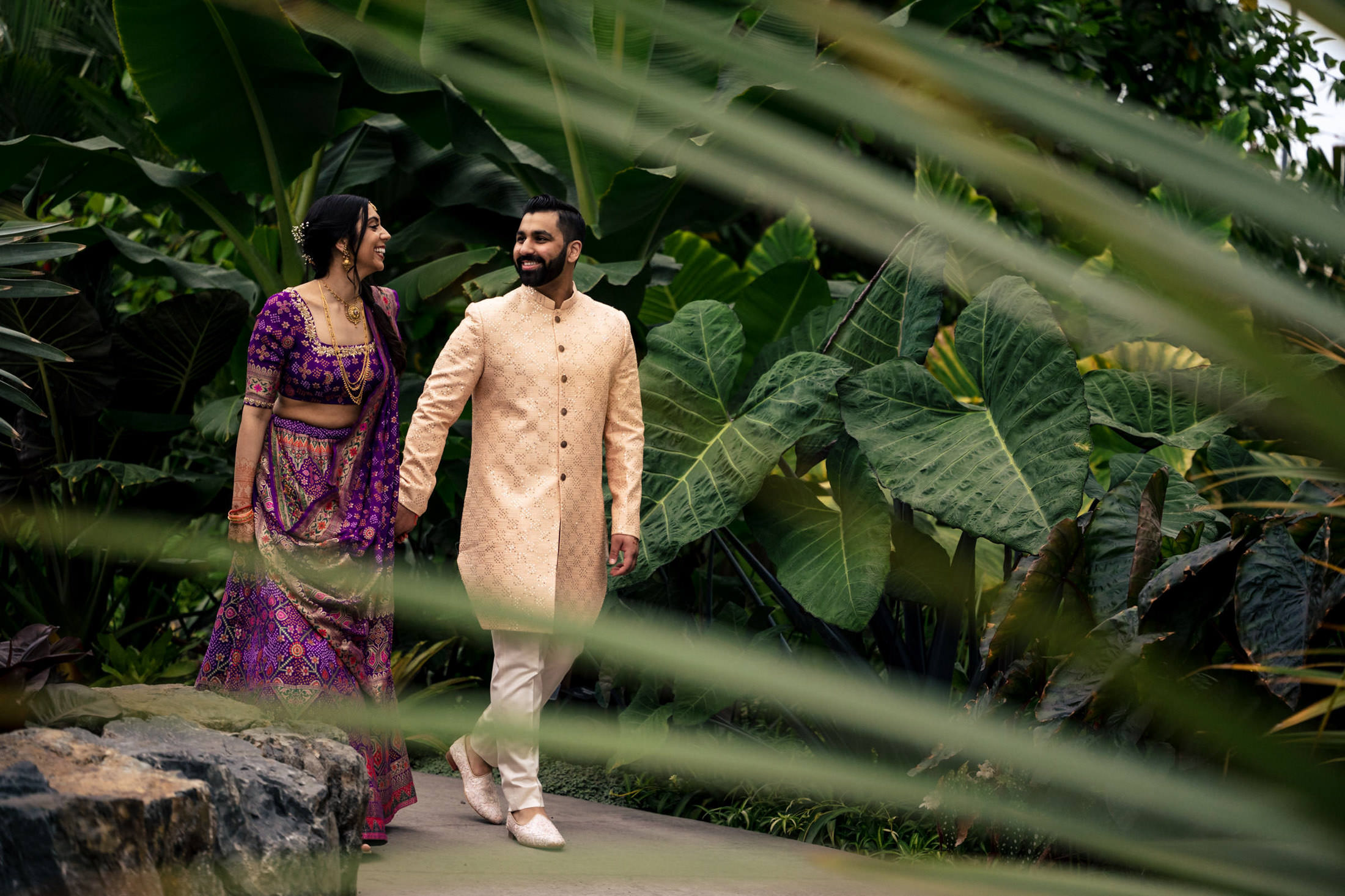 A couple in traditional attire walks joyfully through a lush, tropical garden.