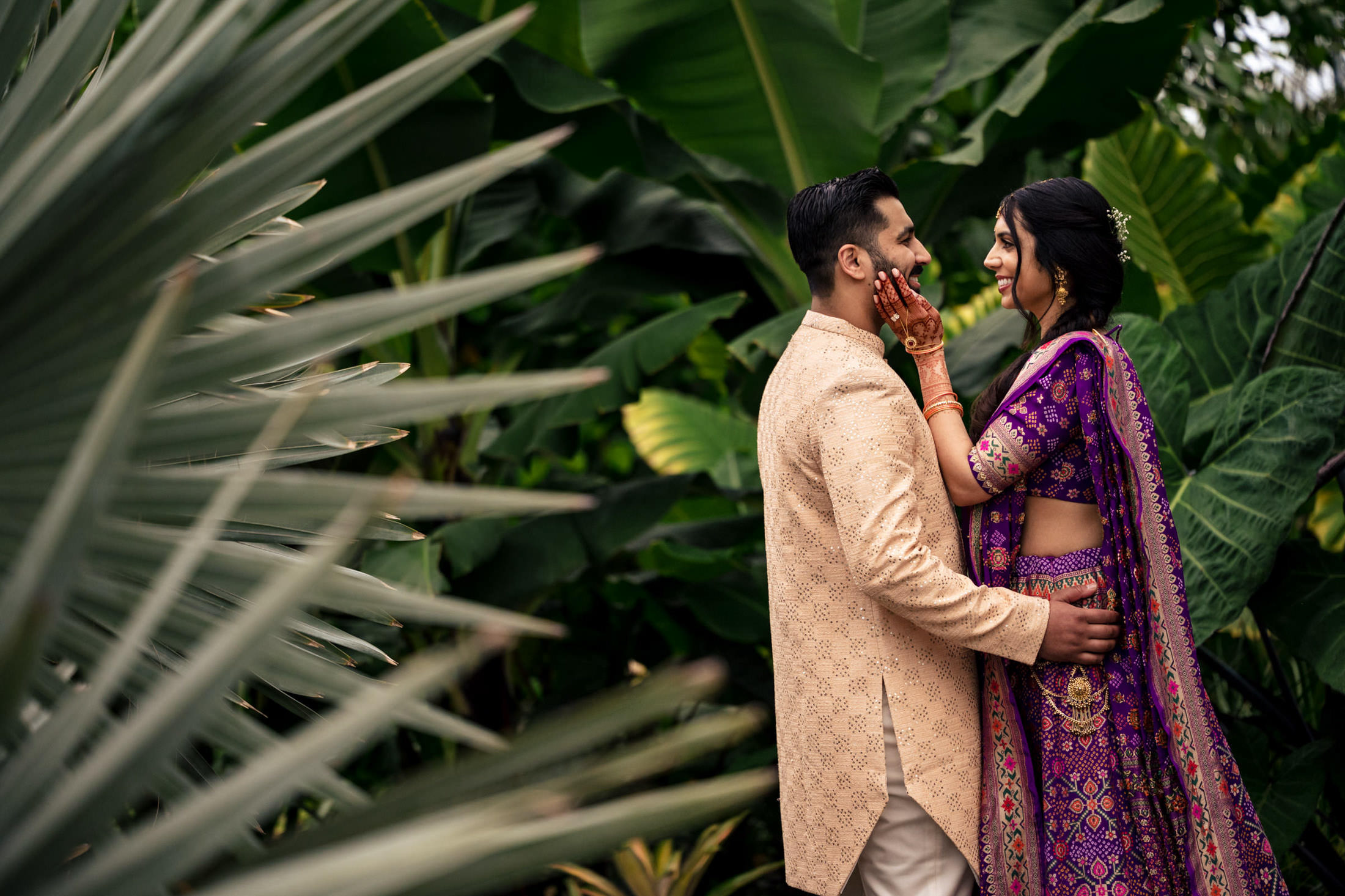 Couple in traditional attire embrace, surrounded by lush green foliage.