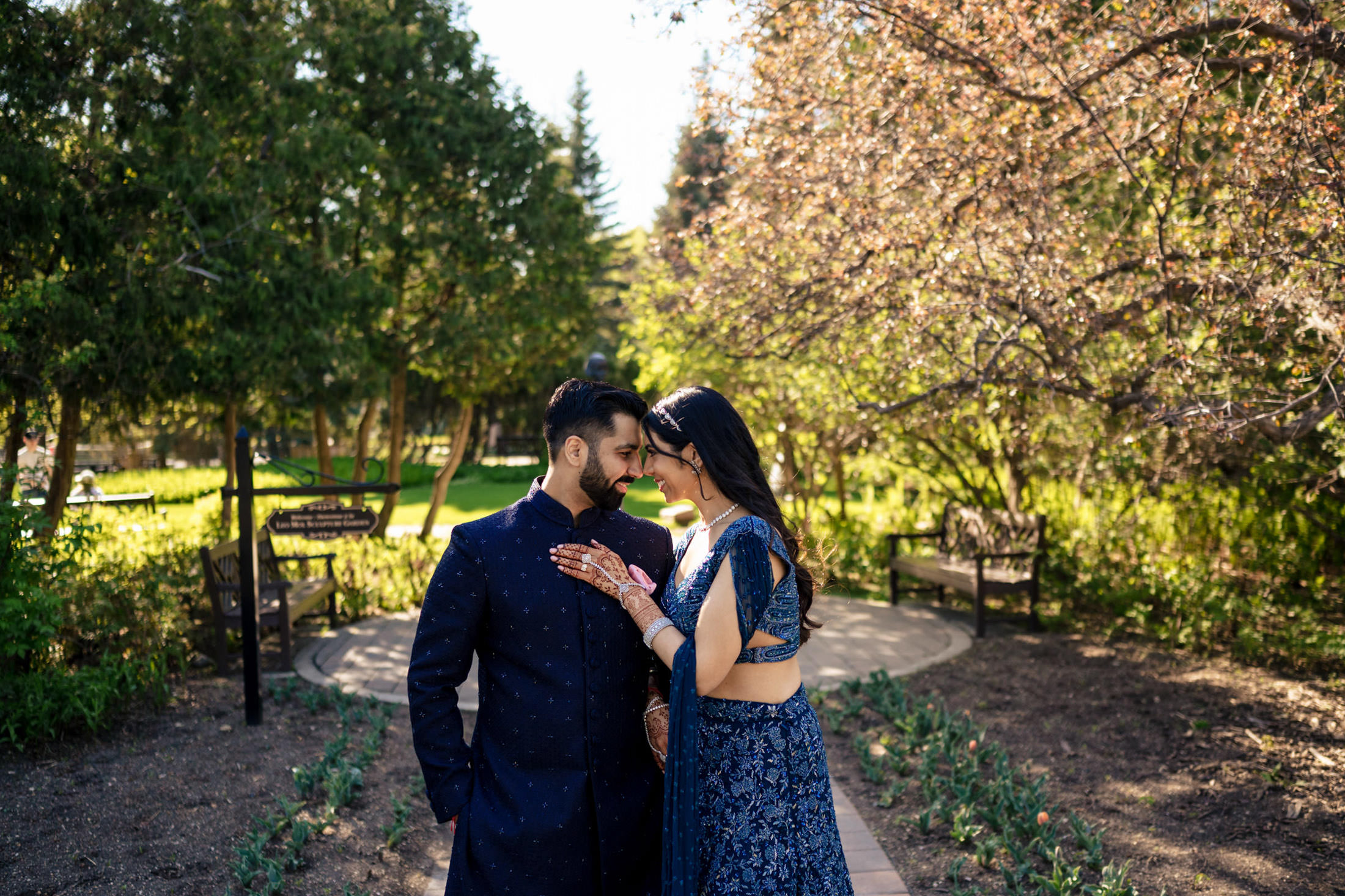 Couple in blue outfits embrace on a garden path under trees.