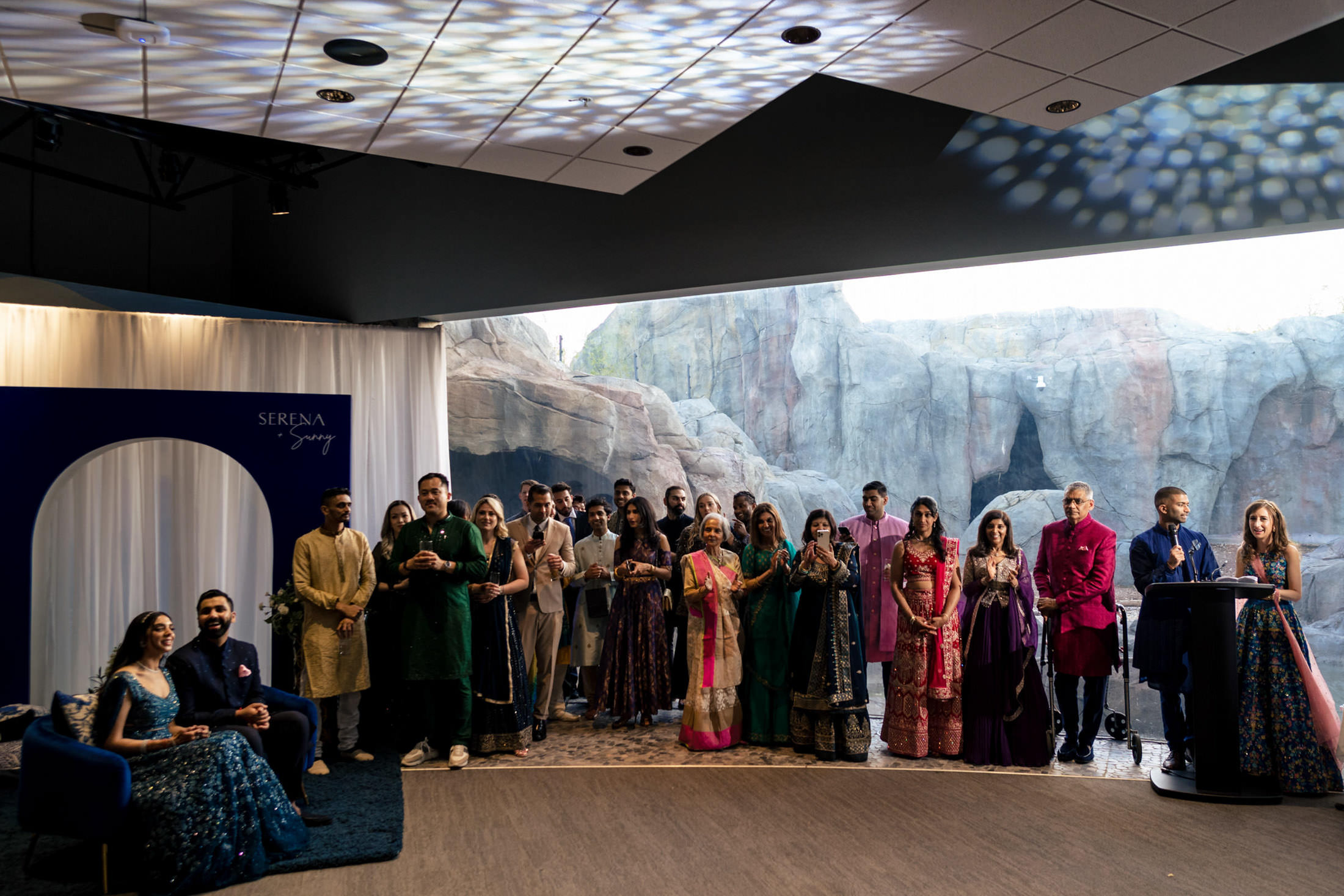 Wedding party in traditional attire standing in front of a rocky backdrop indoors.