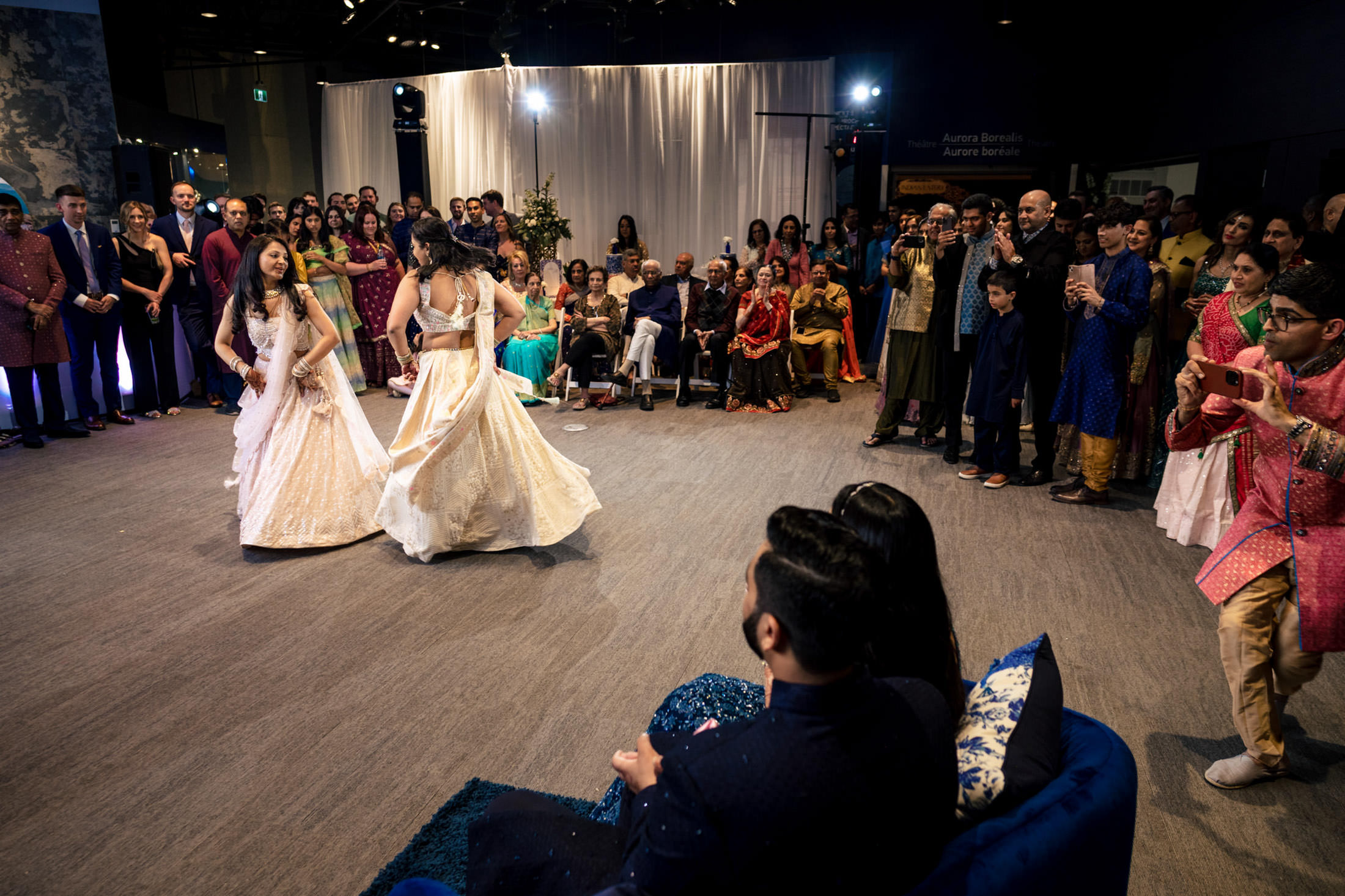 Two women in white dresses dance at an event, surrounded by seated and standing guests.