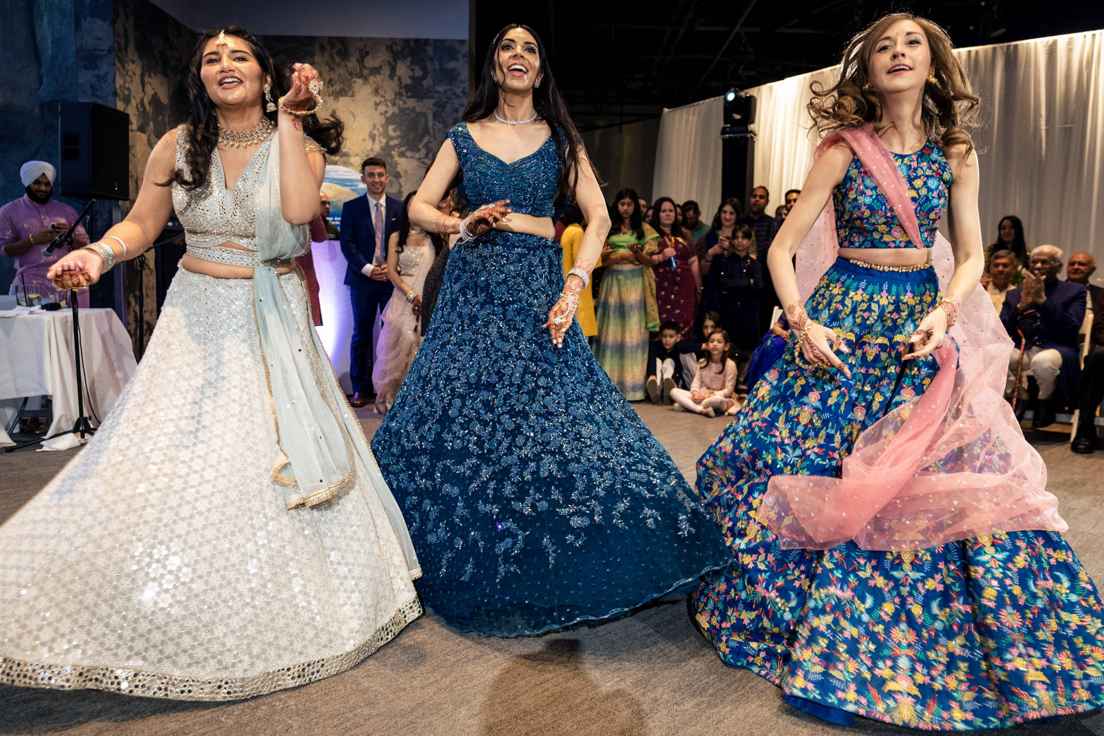 Three women in colorful lehengas dance joyfully at an indoor event with onlookers.