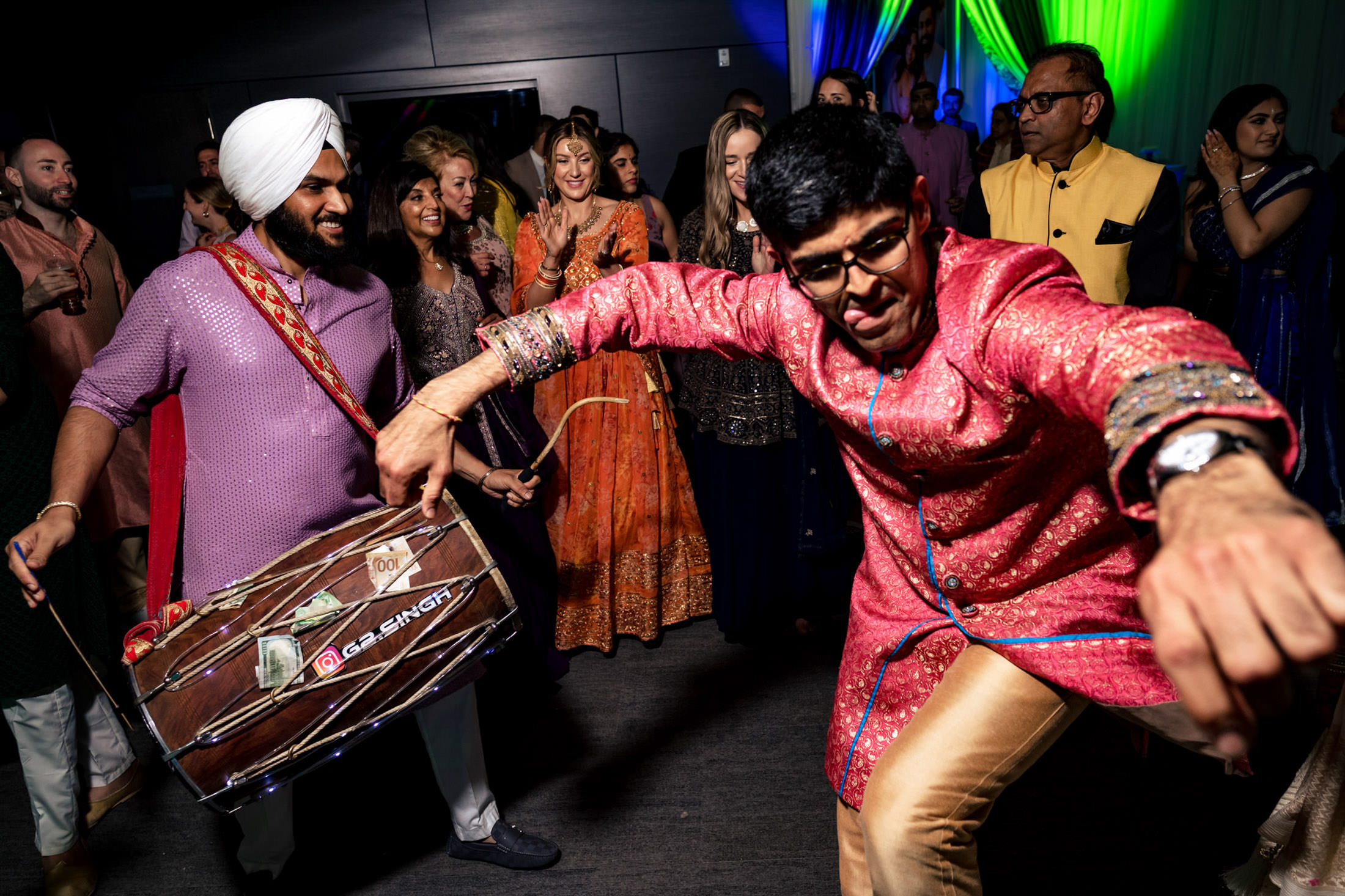 People dancing energetically, one playing a dhol drum at a festive gathering.