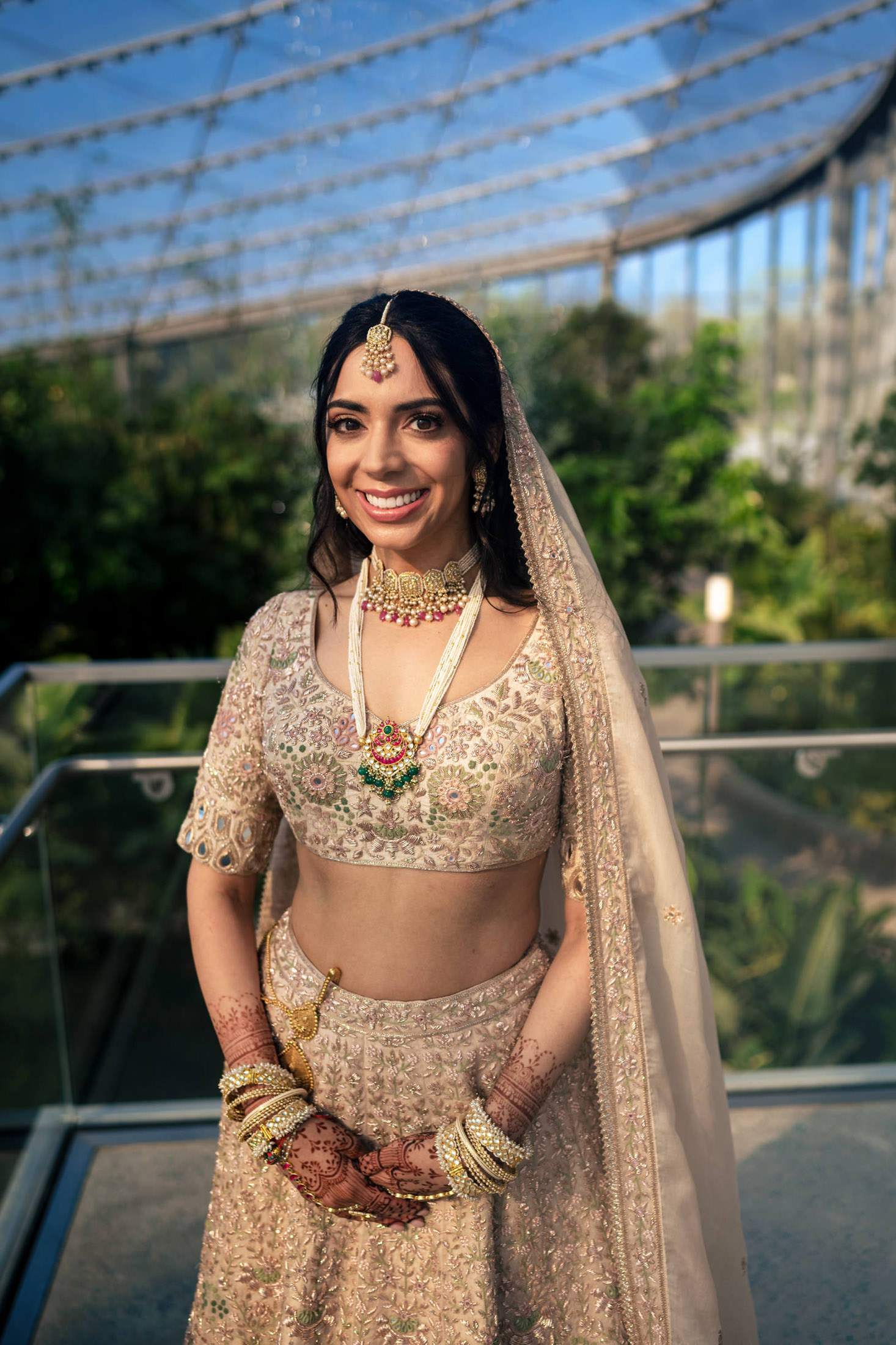 Smiling bride in an ornate lehenga with jewelry, standing outdoors under a glass roof.