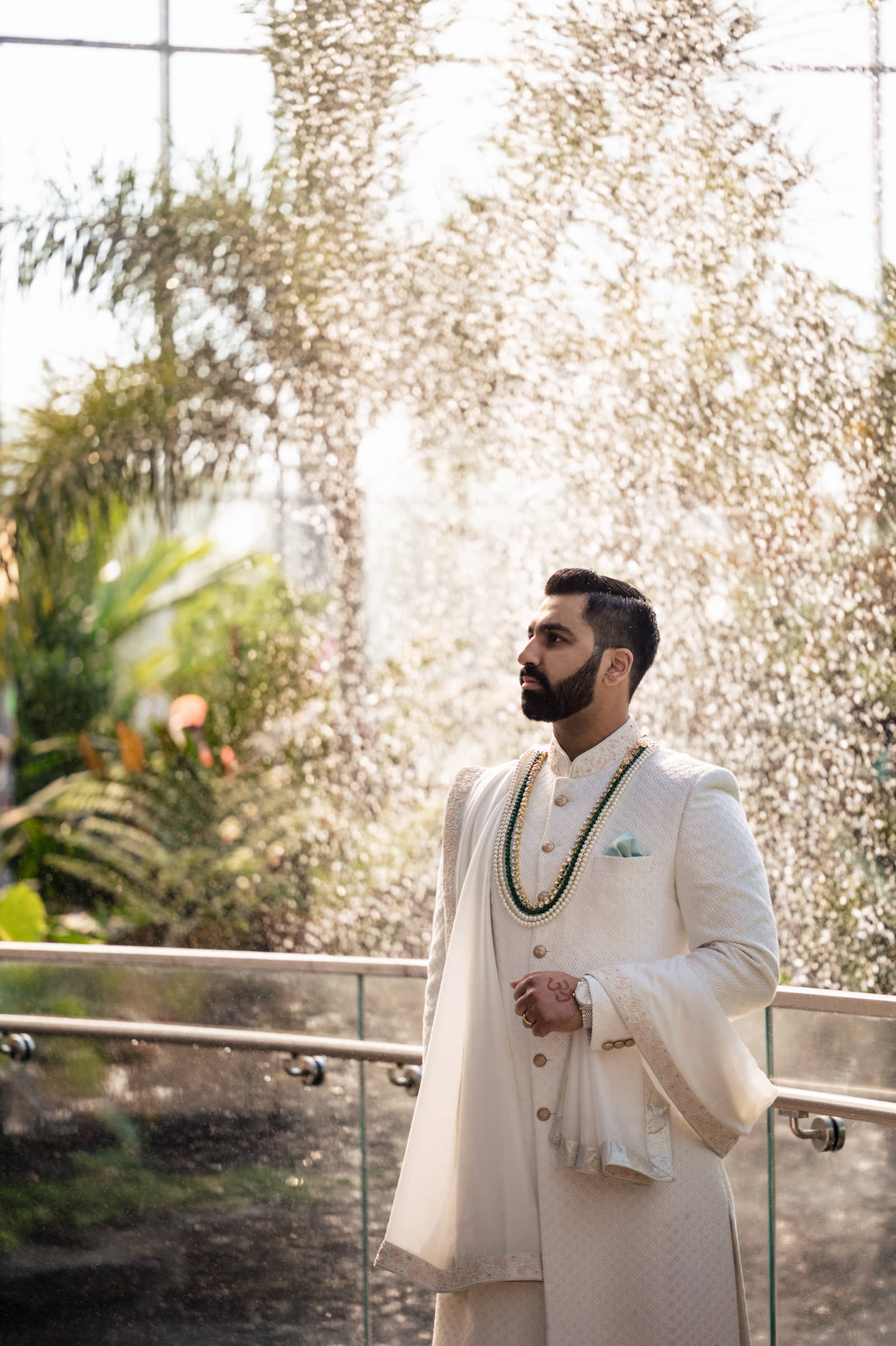 Man in white traditional attire stands before a sparkling water backdrop in a garden.