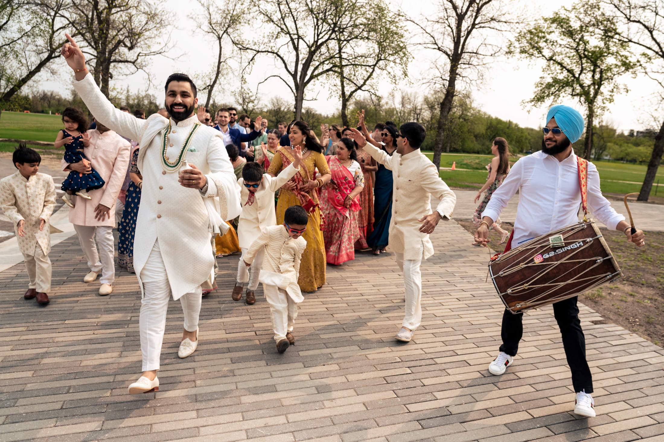 A joyful wedding procession with dancing and a drum player in a park setting.