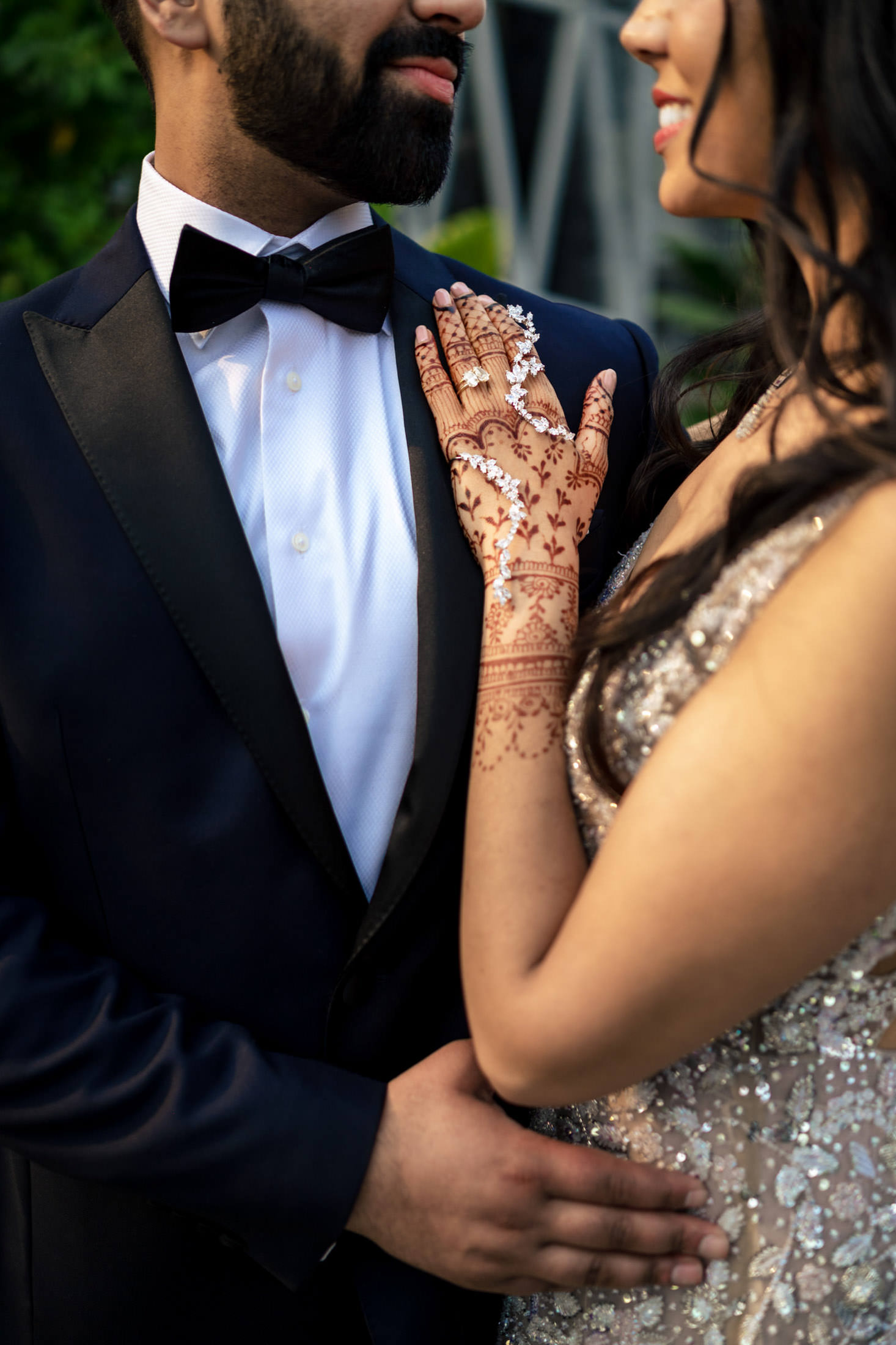 Couple in formal attire; woman with henna on her hand rests it on man's chest.