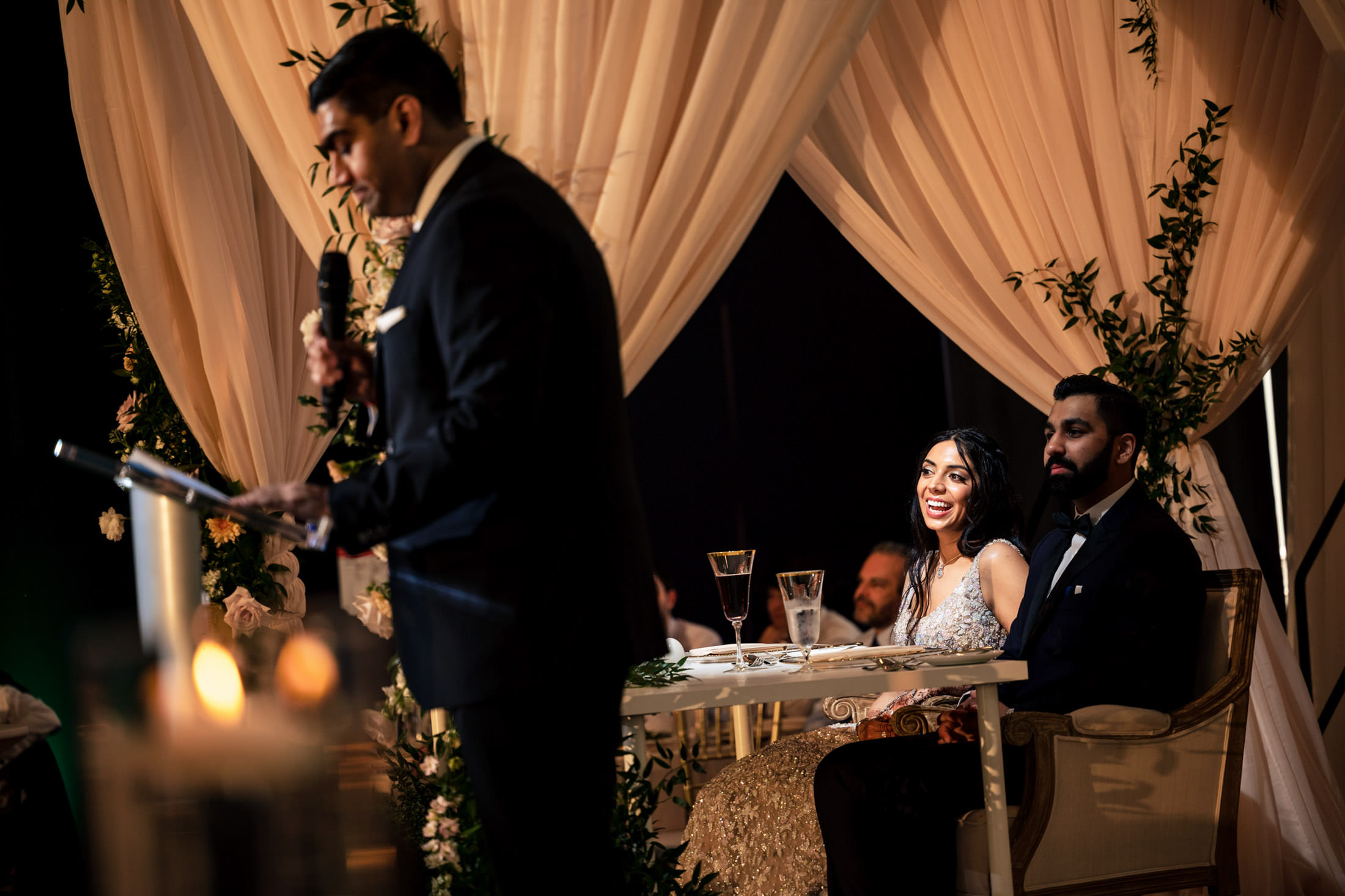 Person giving a speech at a wedding while a couple listens, seated under a floral canopy.