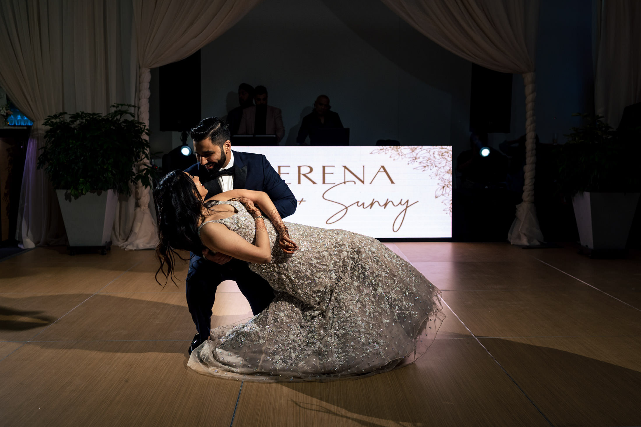 Couple dancing in elegant attire under draped ceiling; backdrop with names displayed.