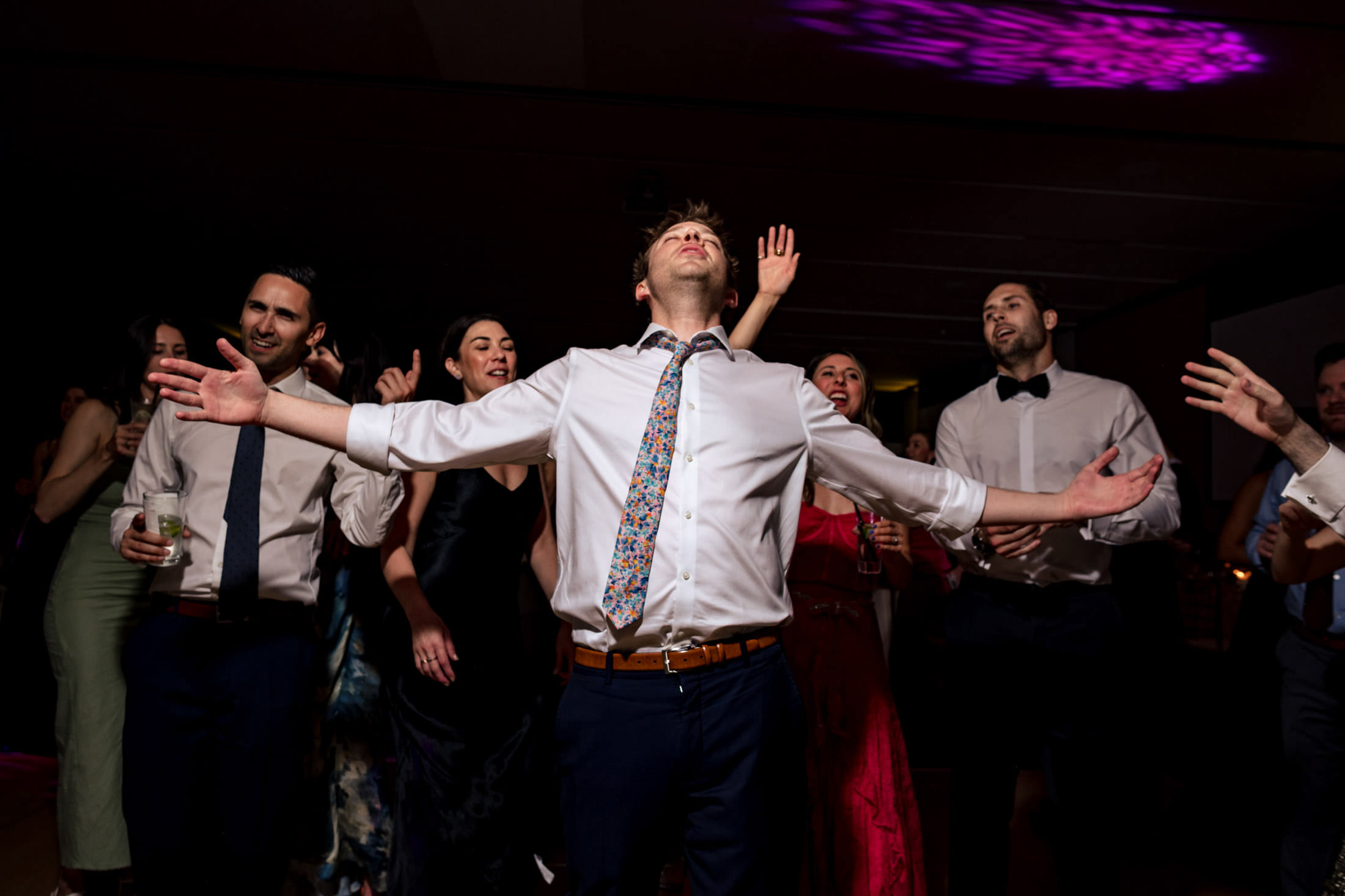 Man with outstretched arms dances joyfully among dressed-up people at an indoor event.