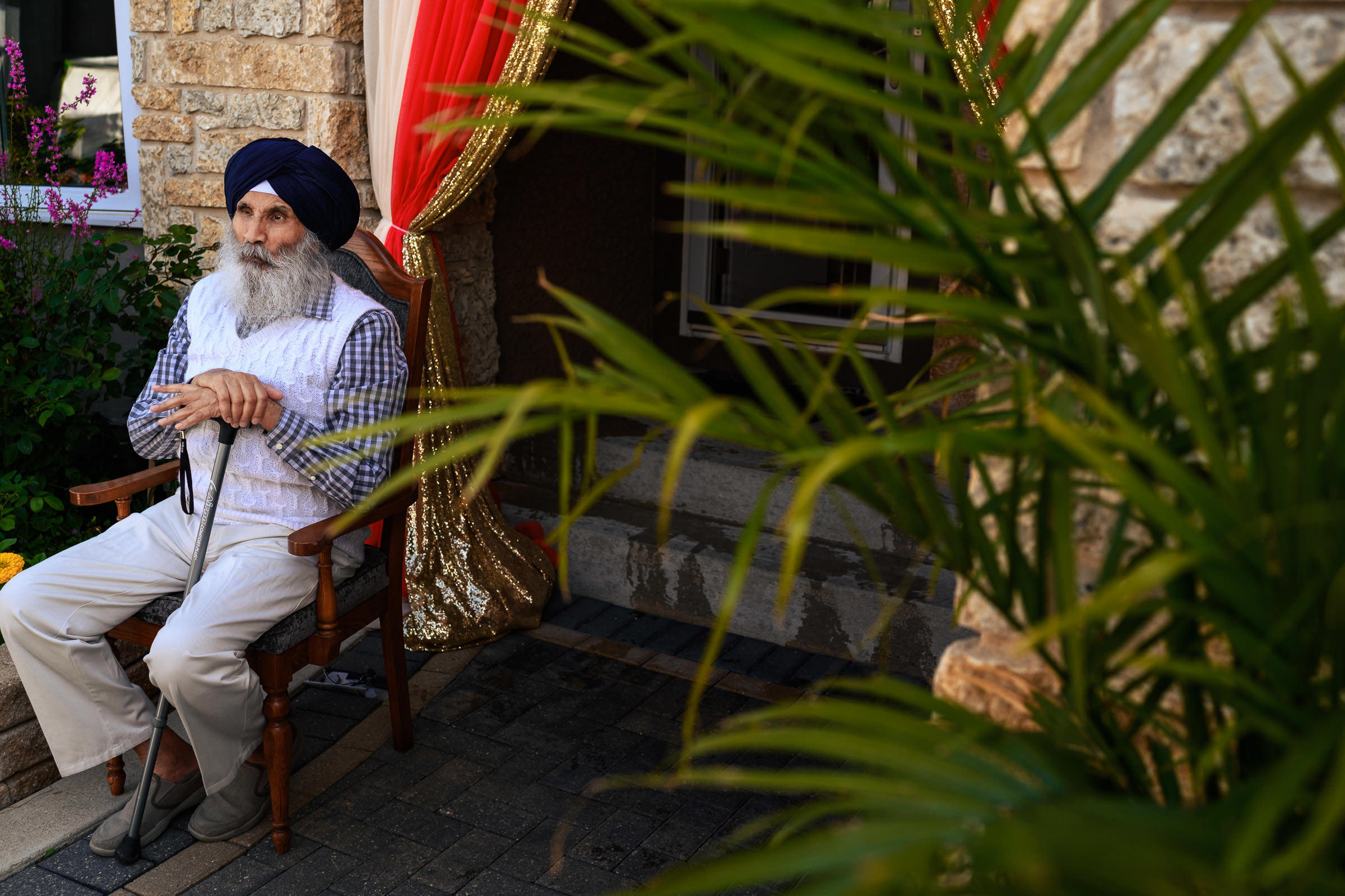 An elderly man in a turban sits outside, framed by red and gold curtains, at an Indian wedding.