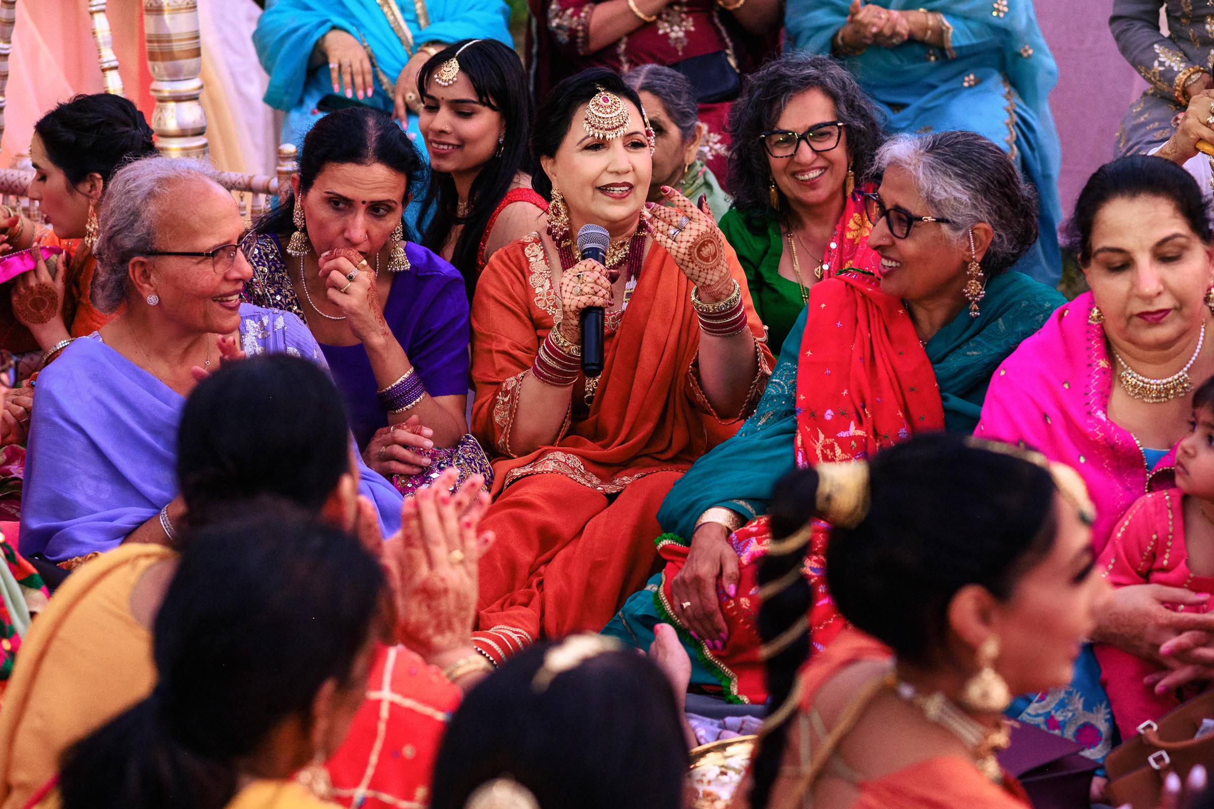 A woman in traditional attire speaks at an Indian wedding, surrounded by colorful clothing.