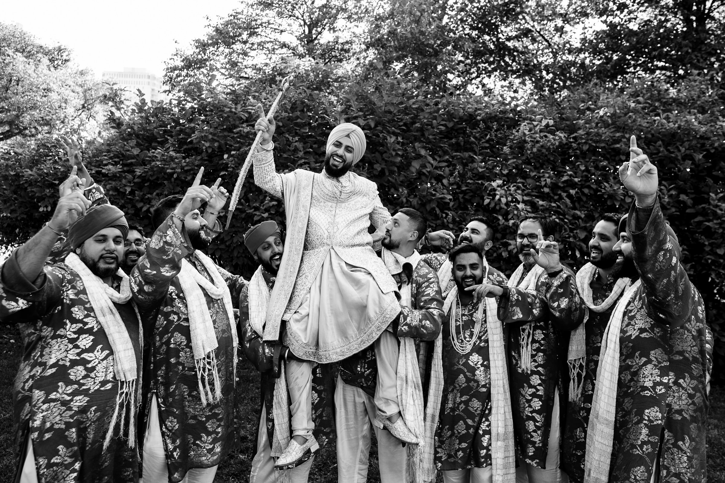 Groomsmen lift a man in traditional attire during an Indian wedding celebration outdoors.