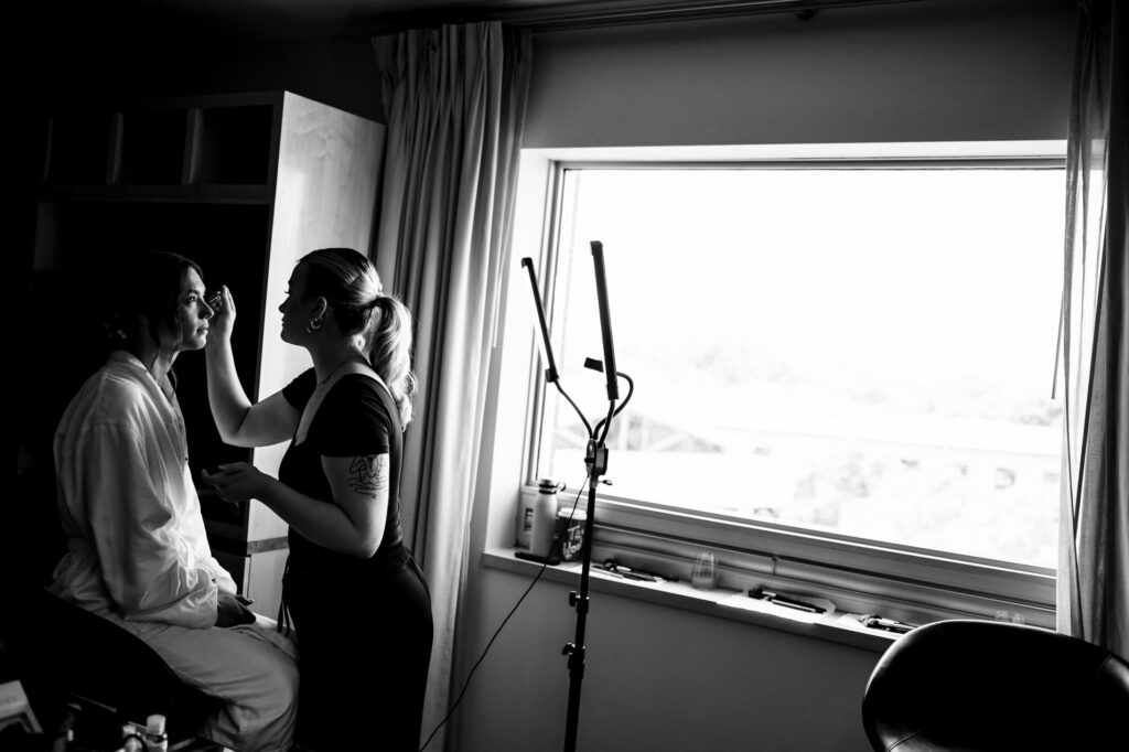 Makeup artist applies makeup to a person sitting near a window in a dimly lit room.